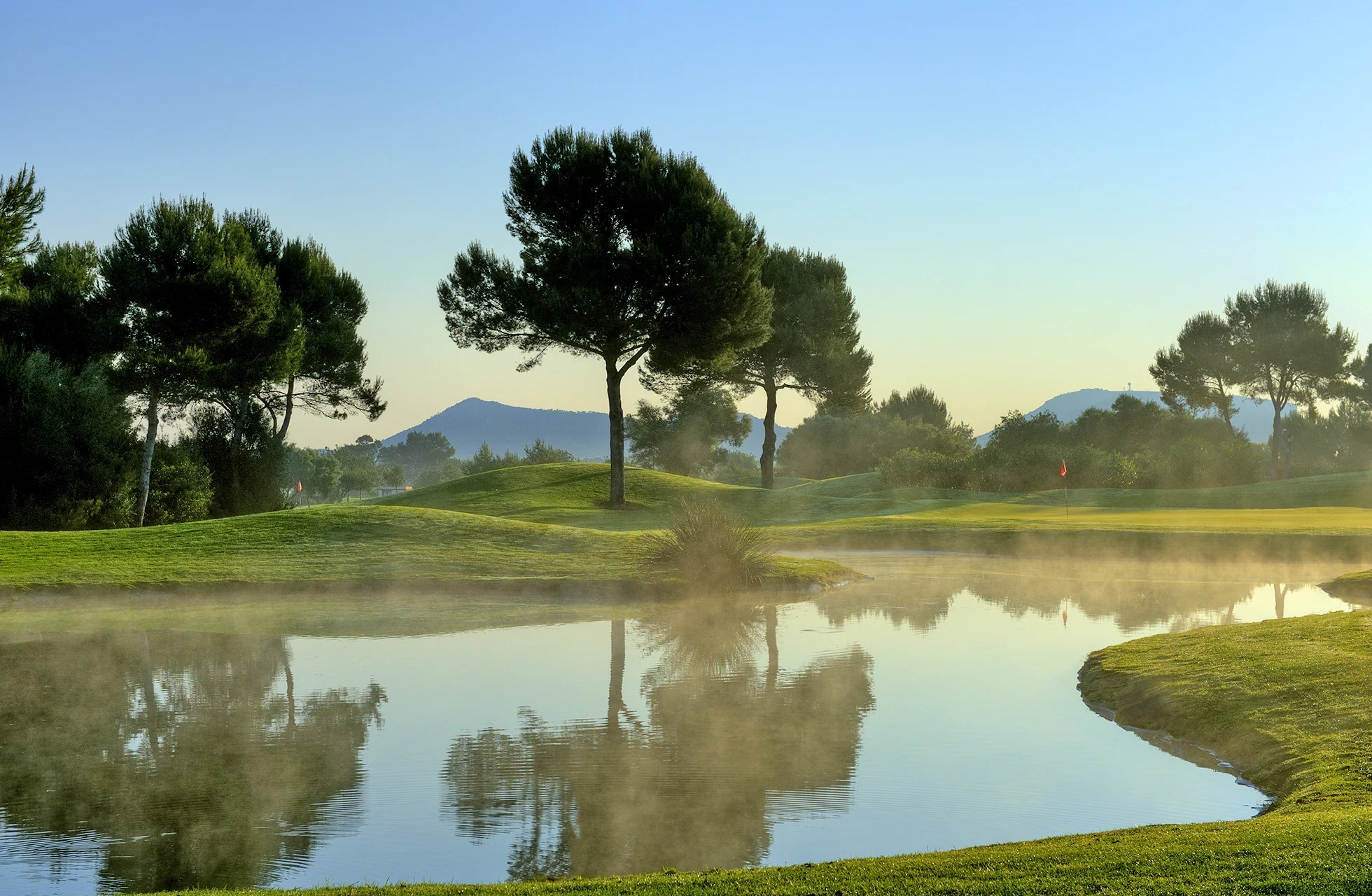 golfbane på son antem golf med vannhinder som speiler trær og himmel, lett dis over vannflaten og fairway i rolig morgenstemning