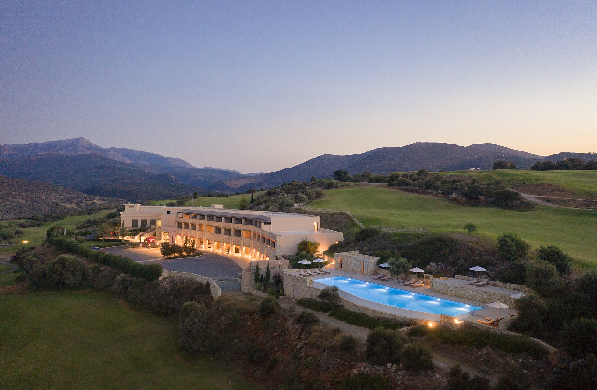 hotellbygning i kveldslys med opplyst basseng, omgitt av golfbane og fjellandskap.