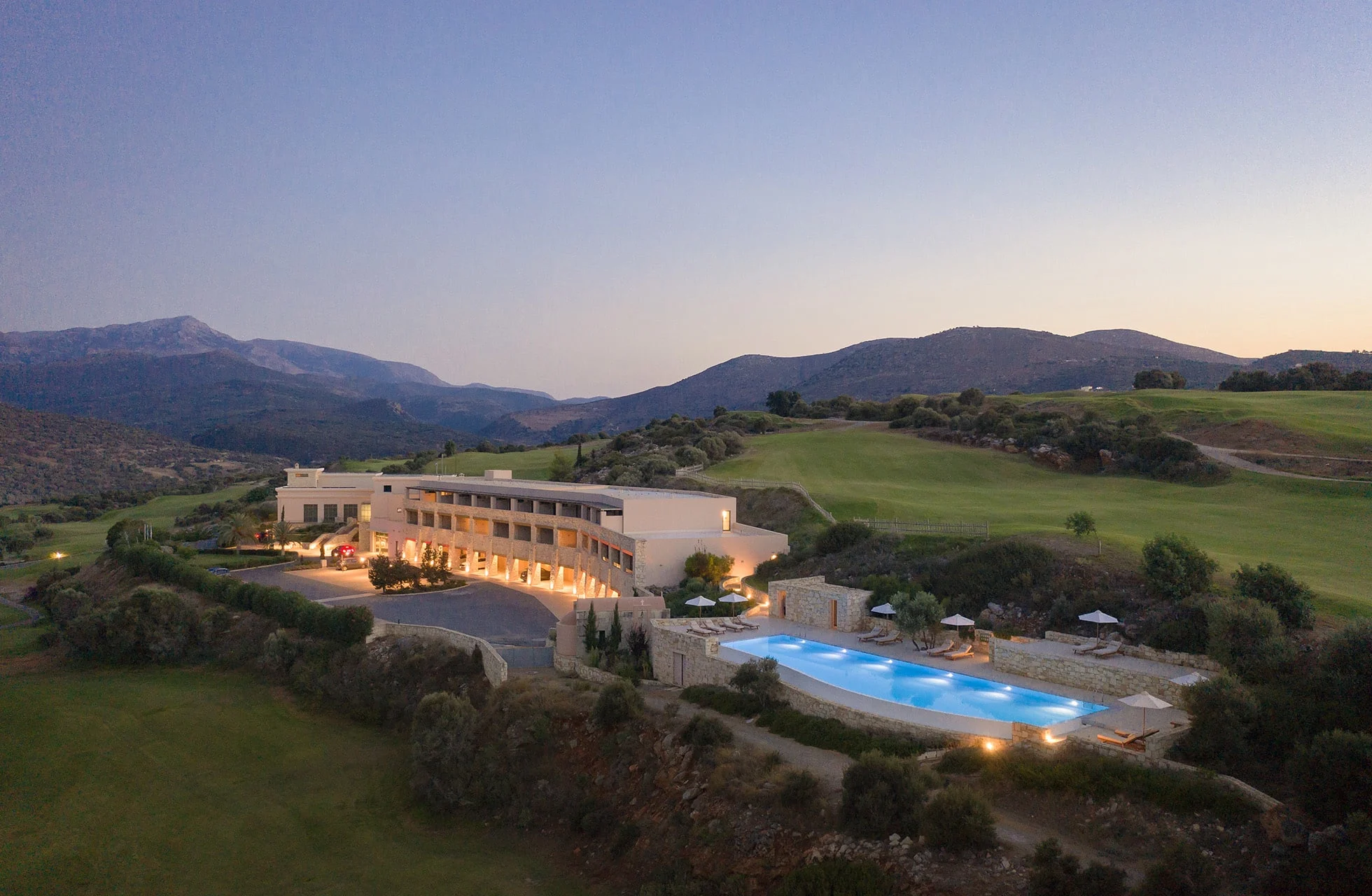 hotellbygning i kveldslys med opplyst basseng, omgitt av golfbane og fjellandskap.