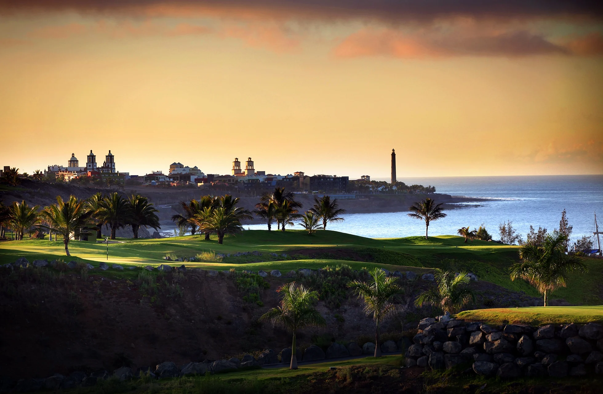 Golfbane i kystlandskab med green og palmer i forgrunden, havet til højre og en by med historiske bygninger og fyr i baggrunden i varmt aftenslys.