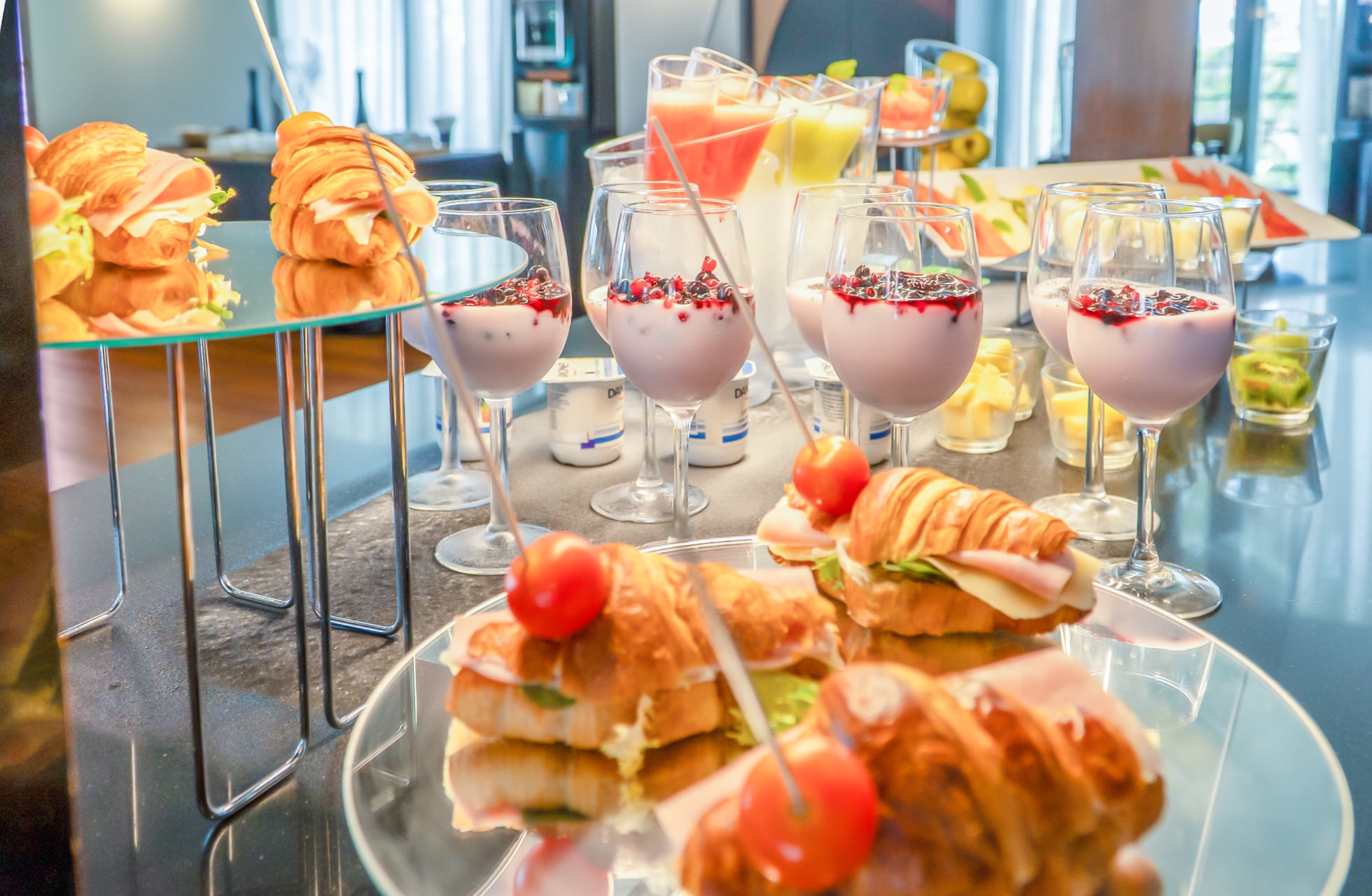 oppdekket buffet med små smørbrød, croissanter og desserter servert i glass, dekorert med frukt og bær på et blankt buffetbord innendørs