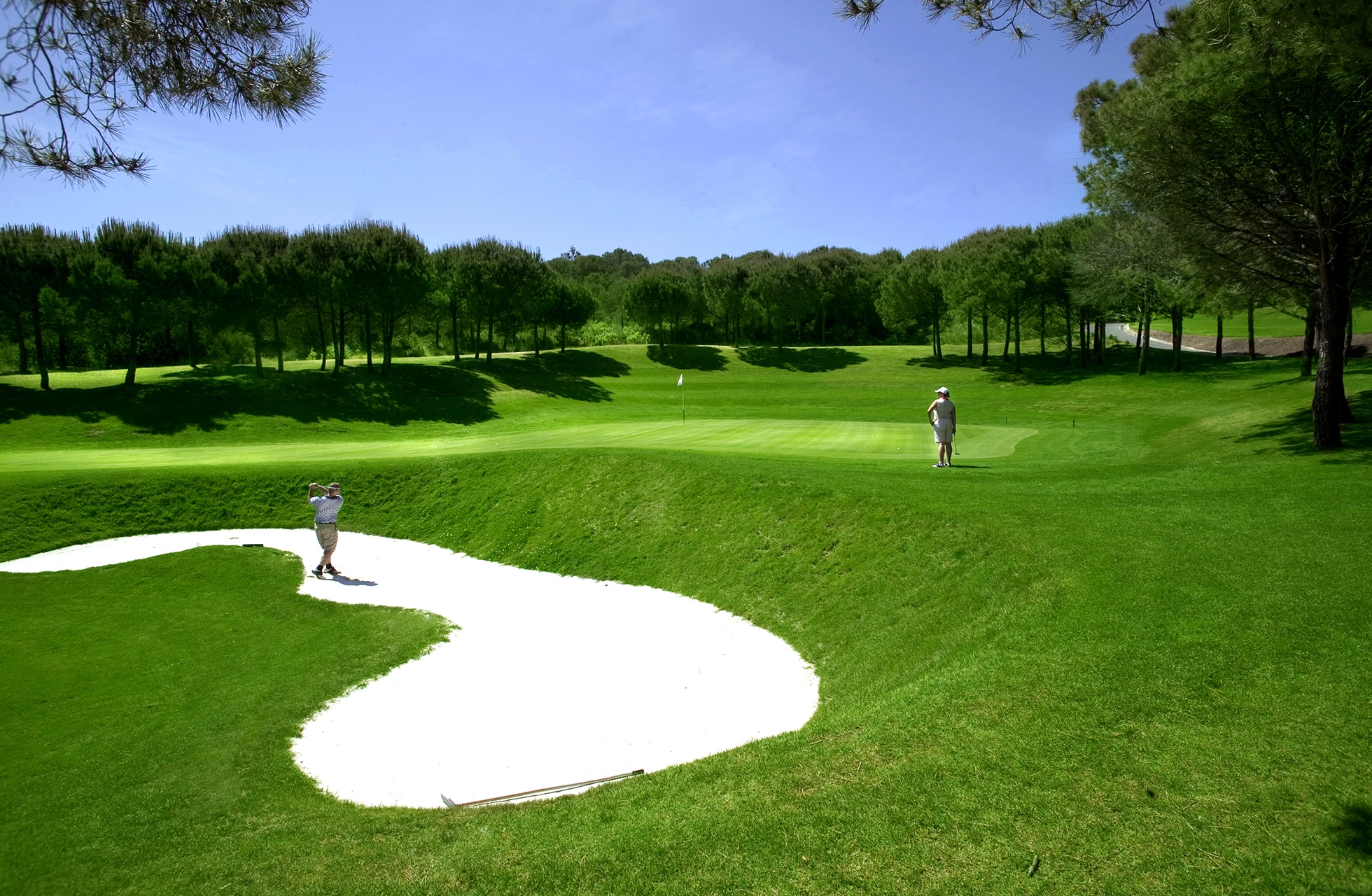 golfbane med sandbunker i forgrunnen, green og fairway i parklandskap samt to golfspillere i klart dagslys