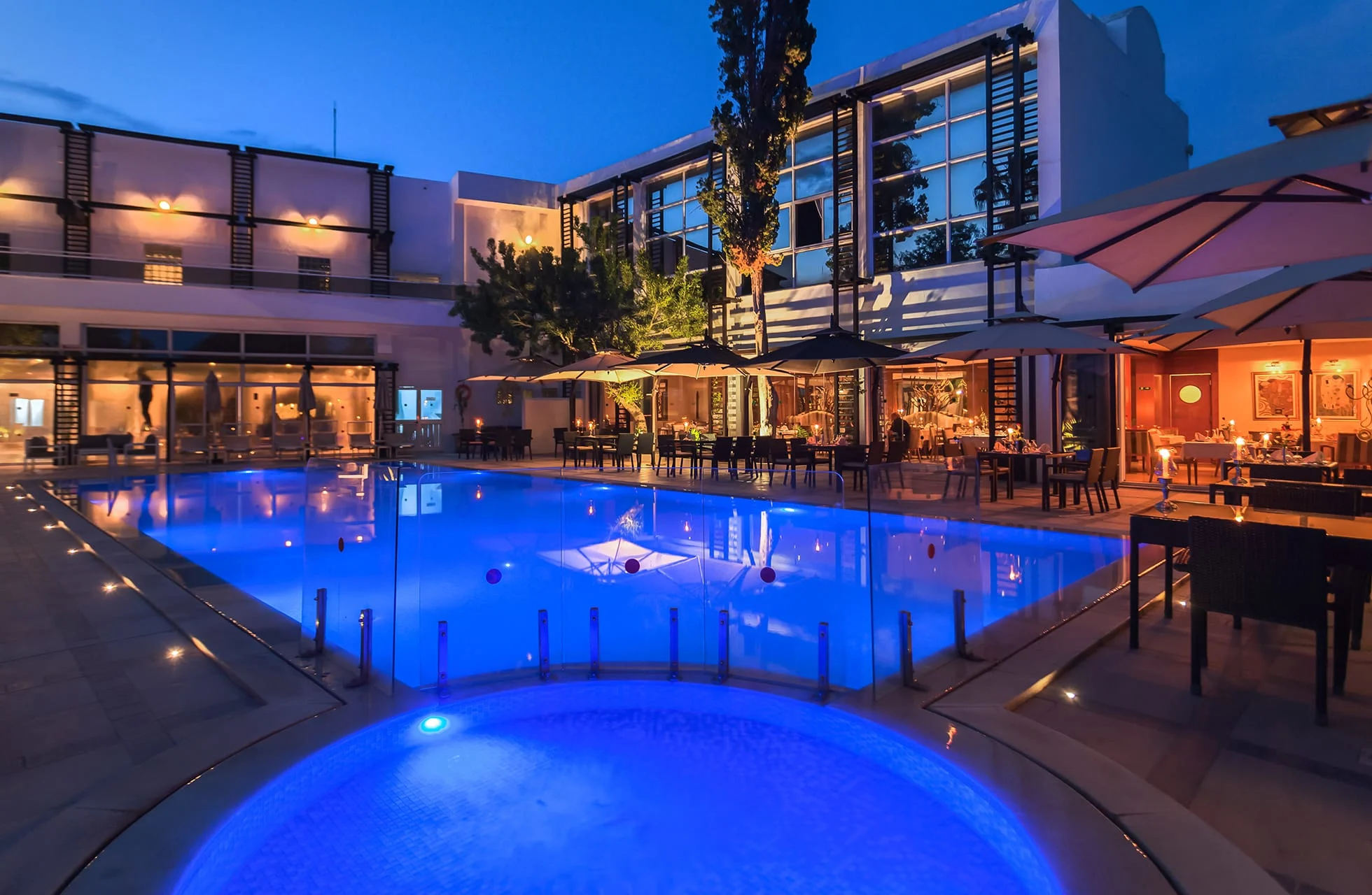 stort opplyst hotellbasseng med avrundet form i forgrunnen, omgitt av solsenger, parasoller og restaurantområder, med hotellbygninger rundt bassengområdet under mørkeblå kveldshimmel