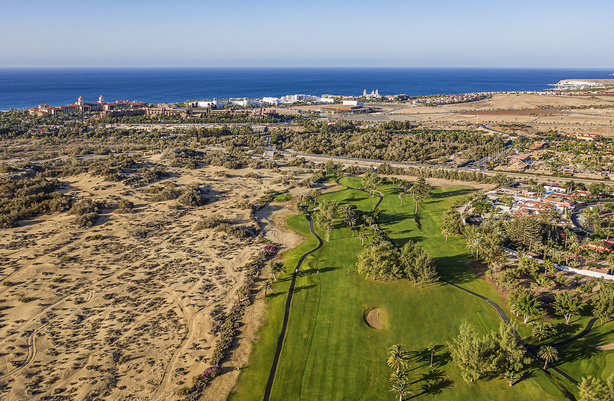 flyfoto av maspalomas golf med grønn fairway og green i kontrast til sanddyner, med havet og kystlinjen i bakgrunnen i klart dagslys