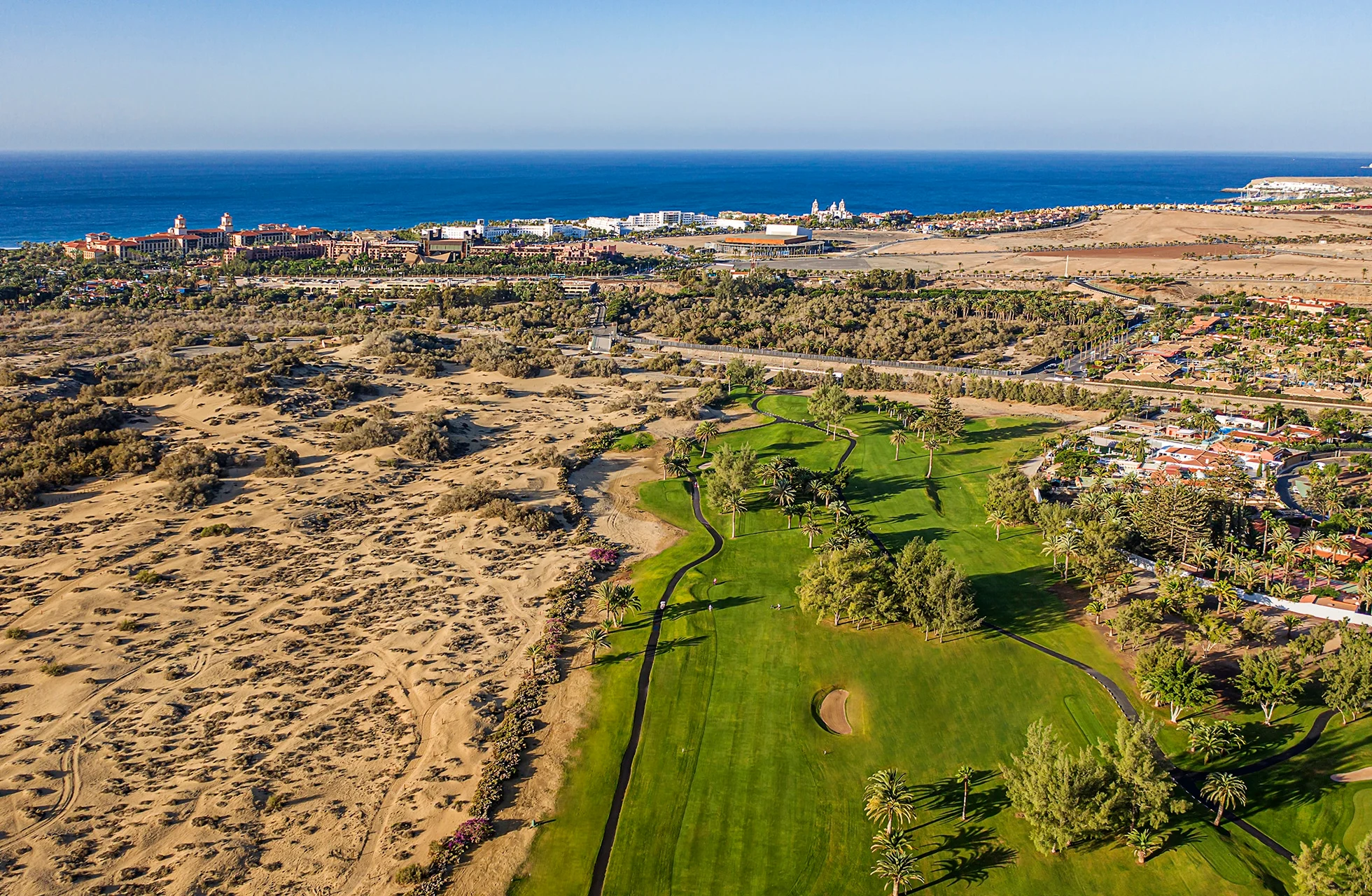 flyfoto av maspalomas golf med grønn fairway og green i kontrast til sanddyner, med havet og kystlinjen i bakgrunnen i klart dagslys