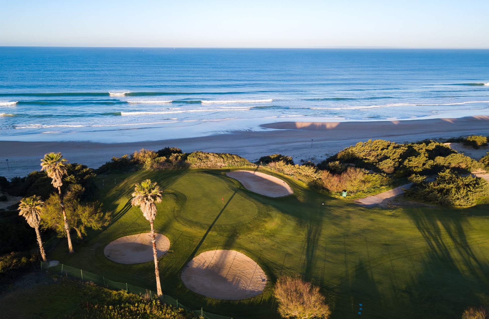 flyfoto av golfgreen med bunkere nær kysten, omgitt av grøntområder og palmer med havet og stranden i bakgrunnen