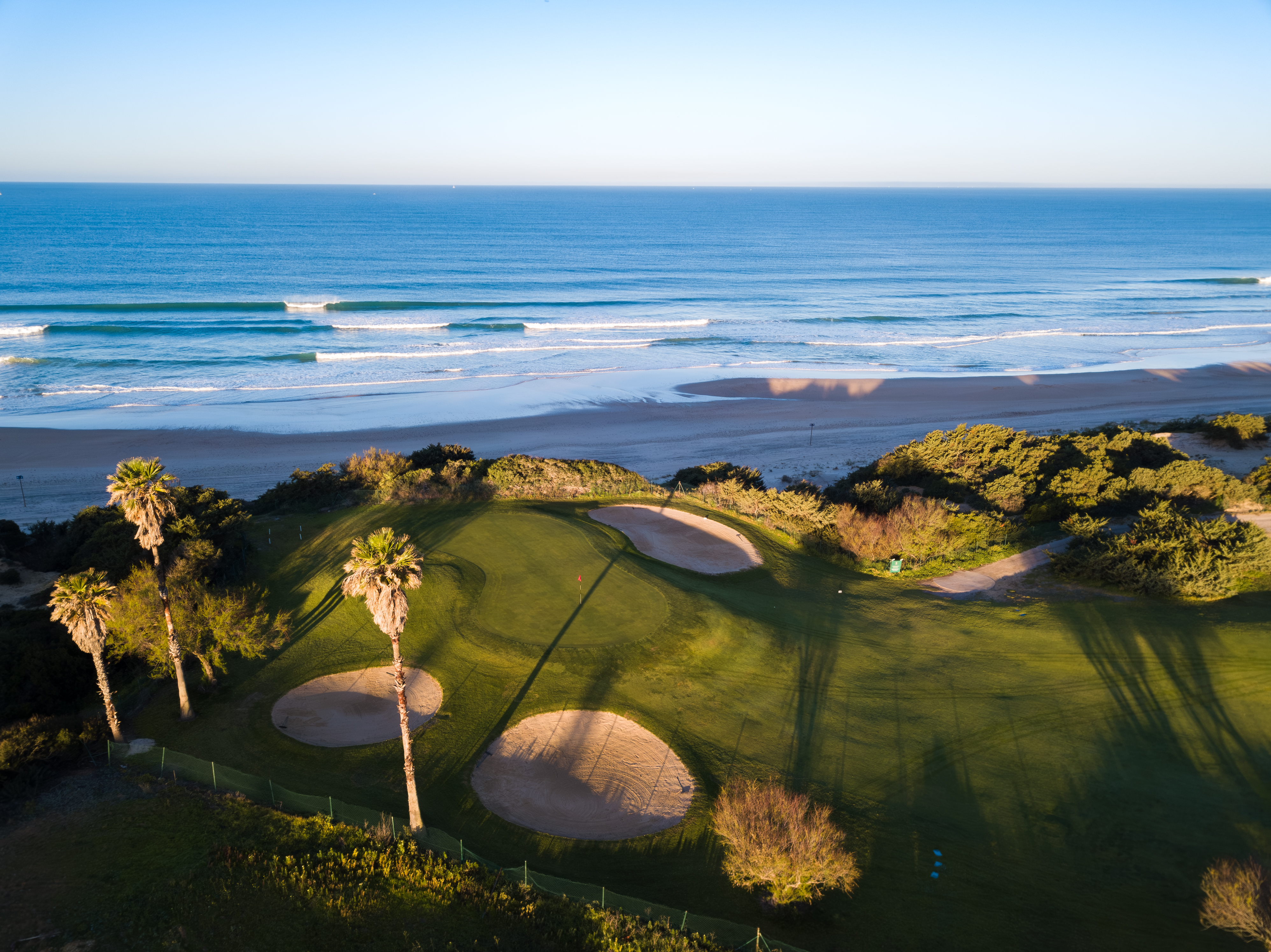 flygvy över golfgreen med bunkrar nära kusten, omgiven av grönska och palmer med havet och stranden i bakgrunden
