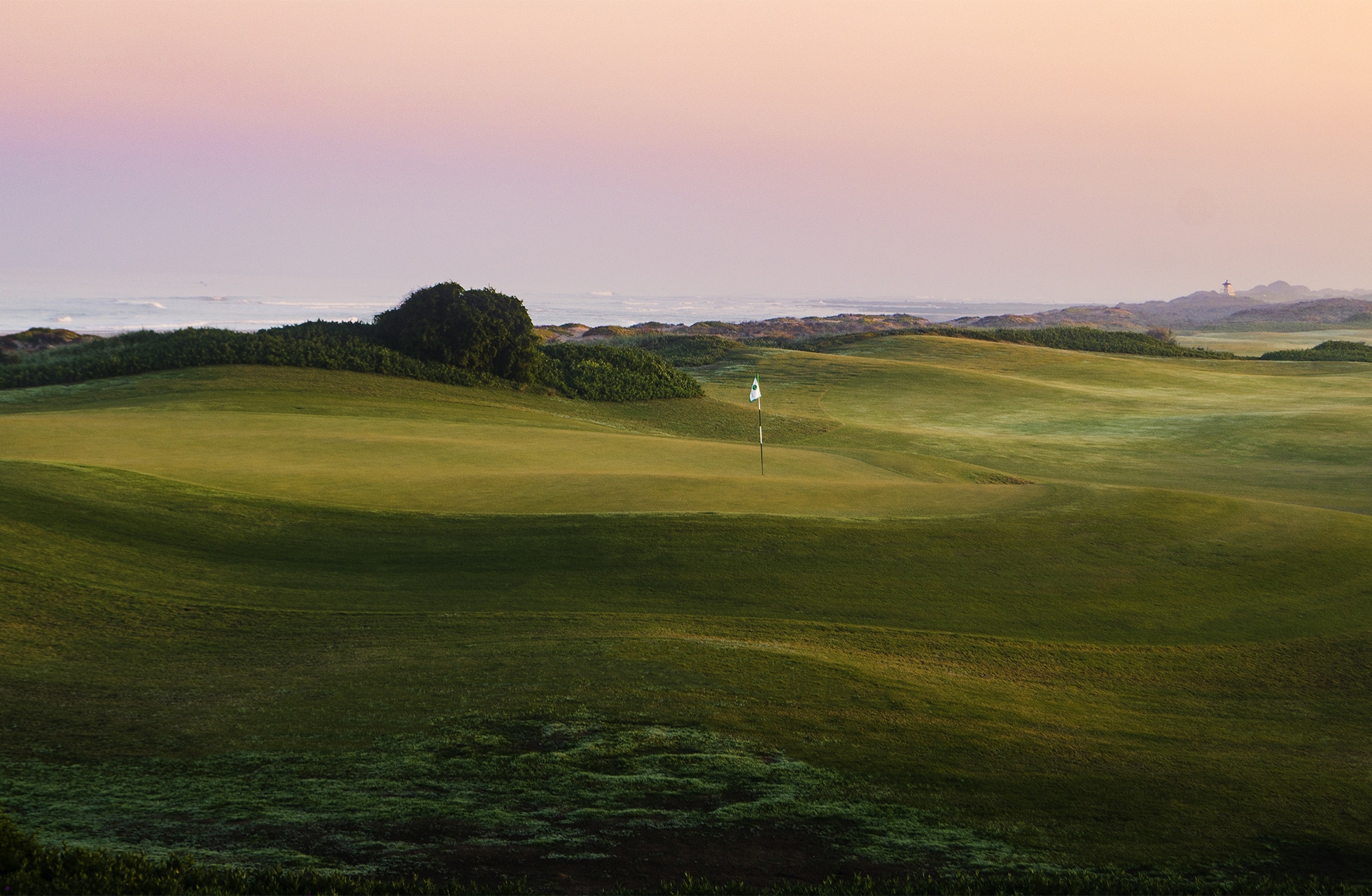golfgreen med flagg på mykt bølgende fairway, omgitt av lav vegetasjon med havet synlig i bakgrunnen i mykt kveldslys