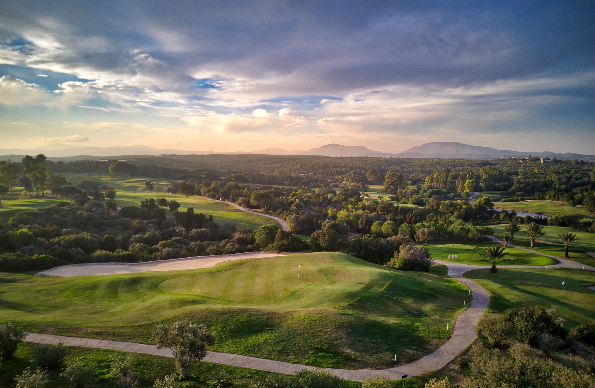 utsikt over en grønn golfbane med fairwayer og greener i kupert landskap, omgitt av trær og med fjell i bakgrunnen under dramatisk himmel