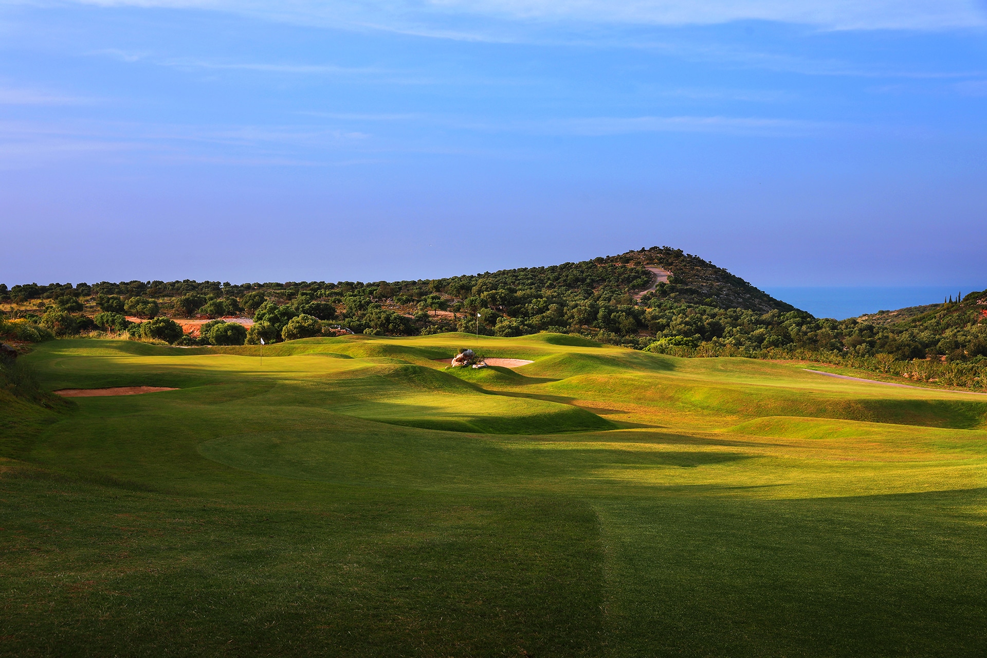 bølgende golfbane med fairwayer og bunkere, grønt kupert landskap og havet synlig i det fjerne.
