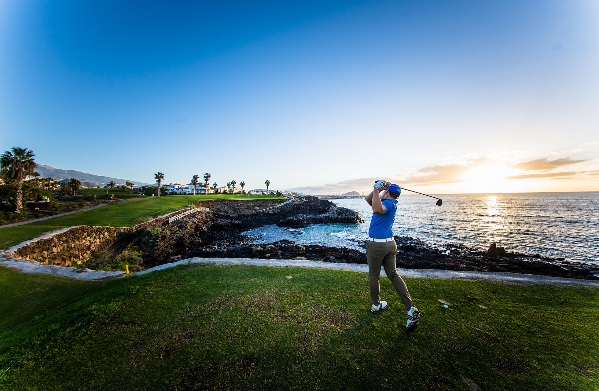 golfaaja lyö golflyöntiä rannikon golfkentällä. viheriö ja väylä sijaitsevat kallioisen rannan vieressä, taustalla meri ja matalalla oleva aurinko lämpimissä sävyissä.