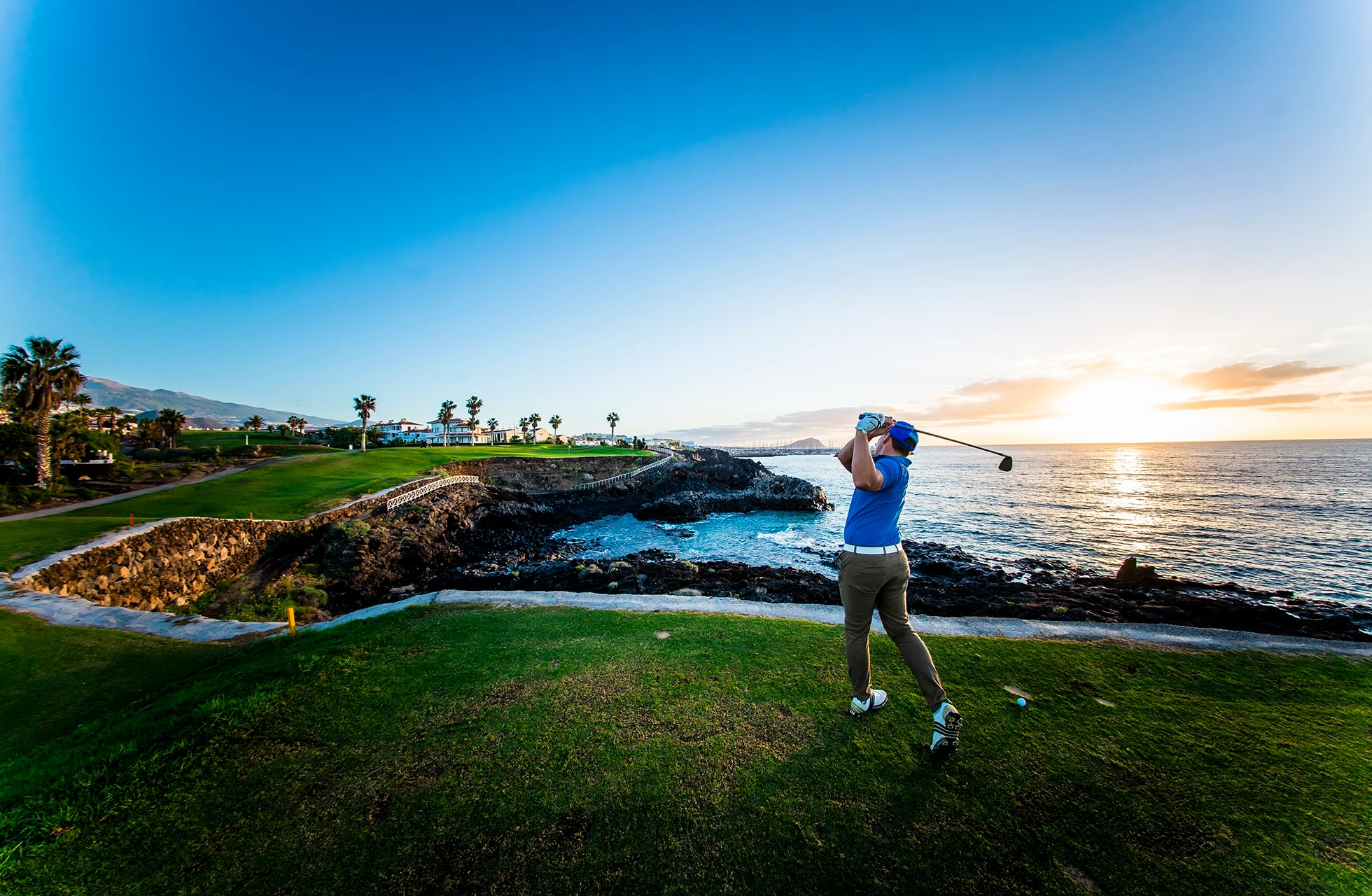 person som slår ett golfslag på en kustnära golfbana. green och fairway ligger intill klippig kust, med havet i bakgrunden och låg sol som färgar himlen i varma toner.