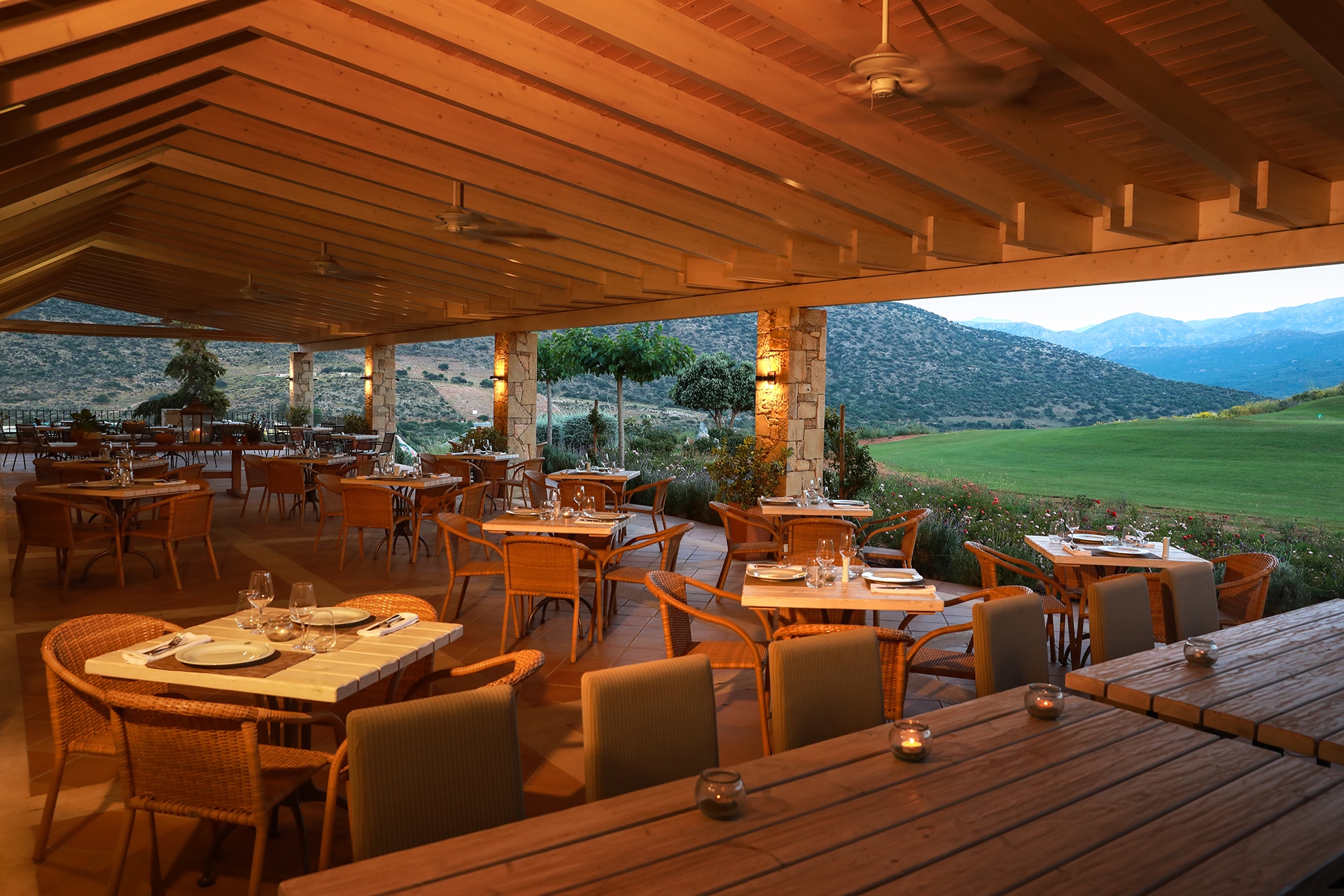 overbygd utendørsrestaurant med dekkede bord og varm belysning, med utsikt over grønt landskap og fjell ved en golfbane