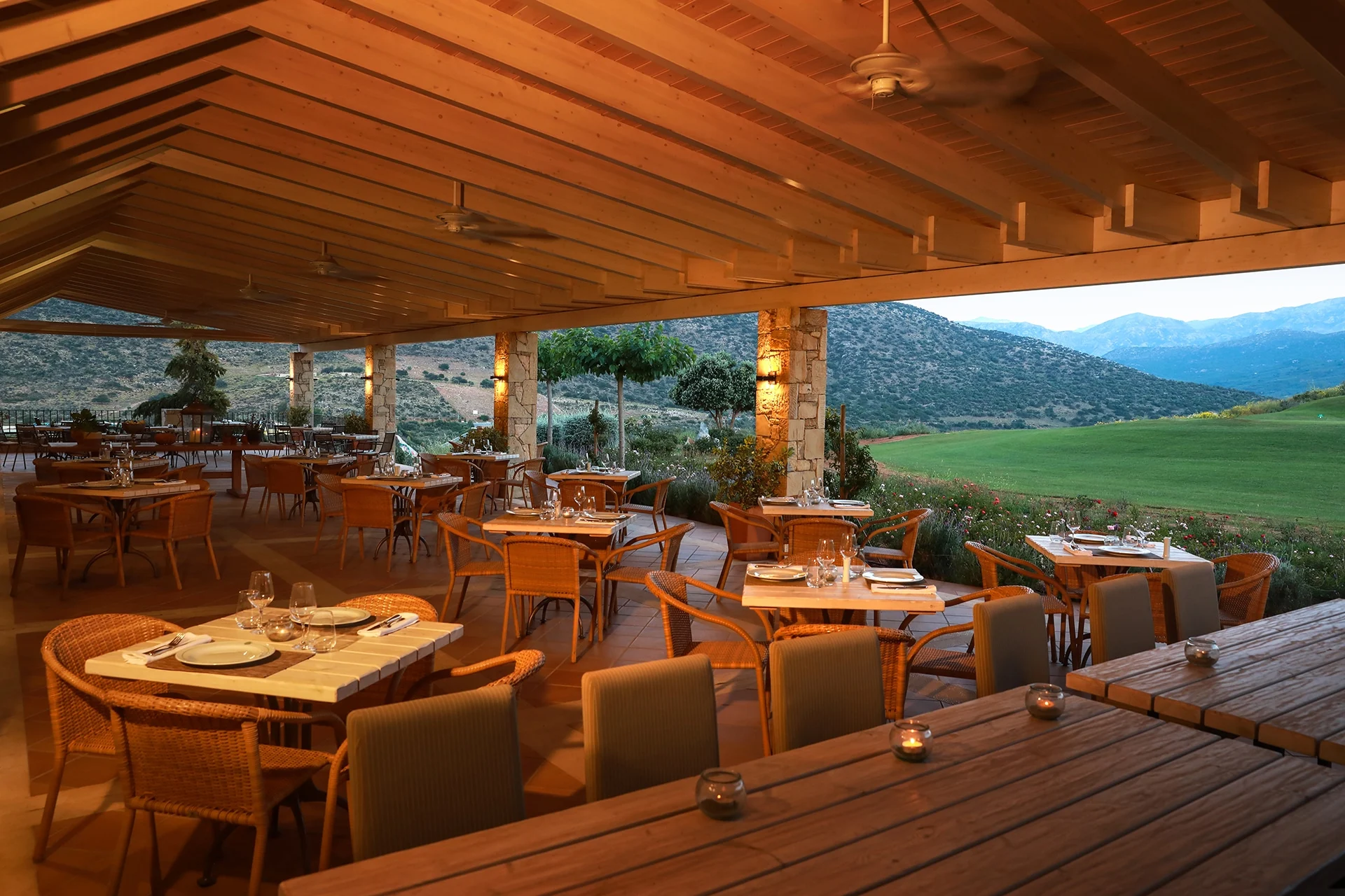overbygd utendørsrestaurant med dekkede bord og varm belysning, med utsikt over grønt landskap og fjell ved en golfbane