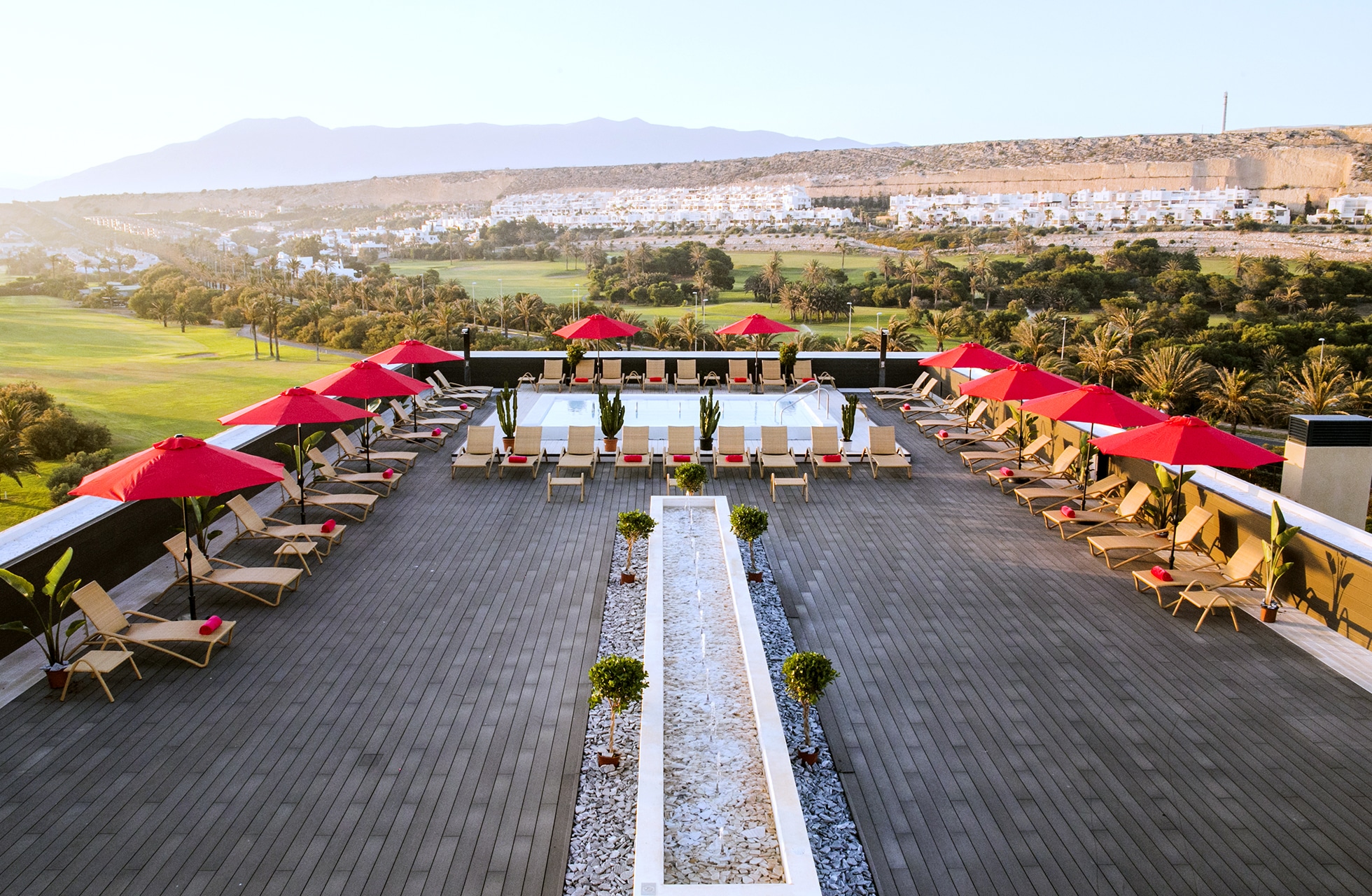 hotellterrasse med basseng, røde parasoller og solsenger med utsikt over golfbanen