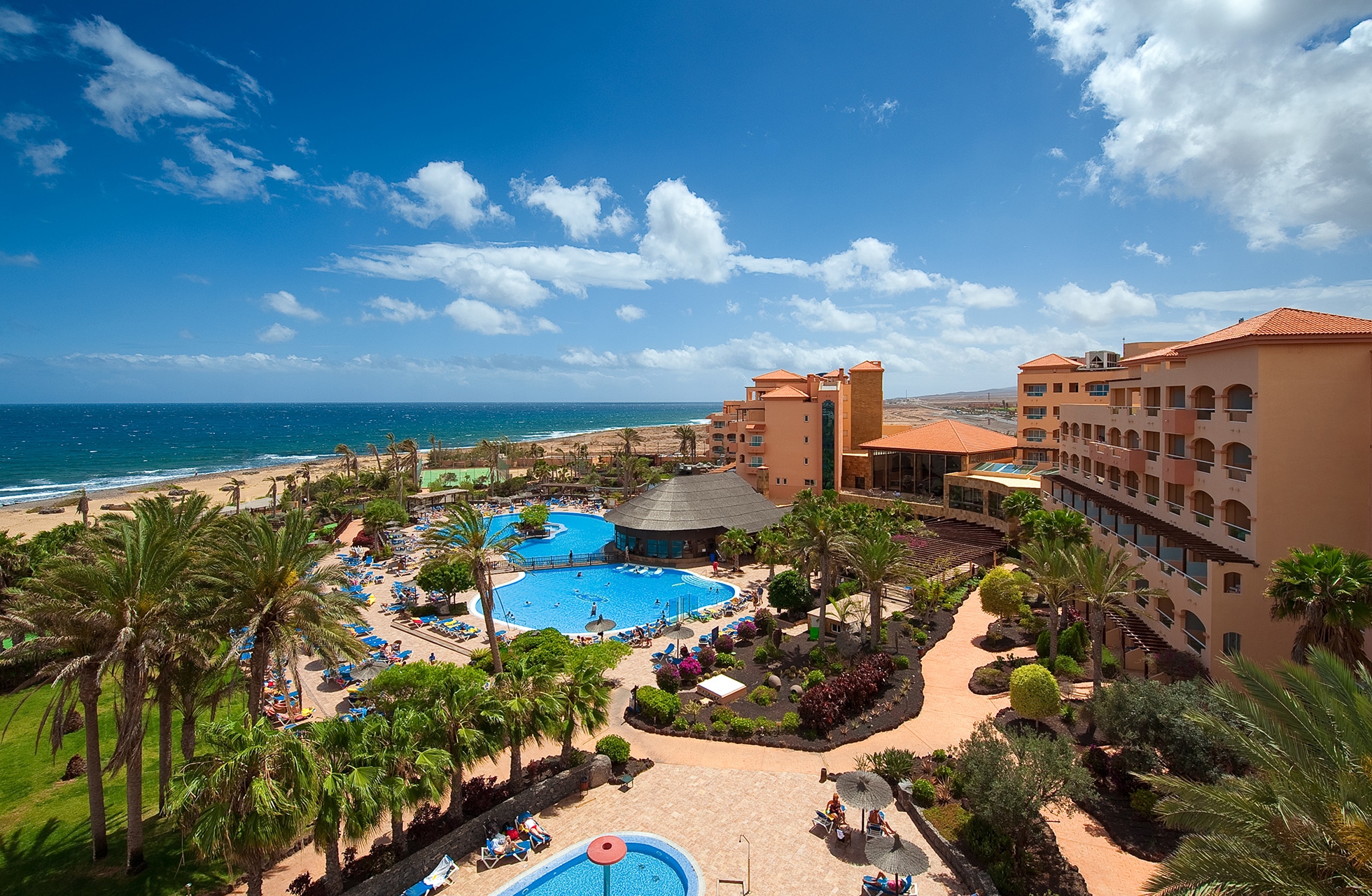 panoramautsikt over kystnært hotellresort med flere bassenger, palmer, hotellbygninger og havet i bakgrunnen i klart dagslys