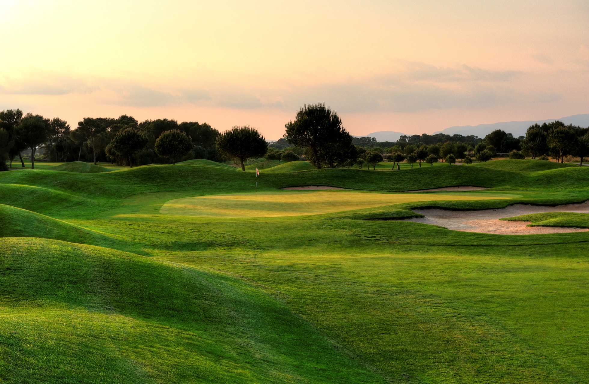 golfgreen på son antem golf med bunker i forgrunnen, omgitt av bølgende fairwayer og trær i varmt kveldslys