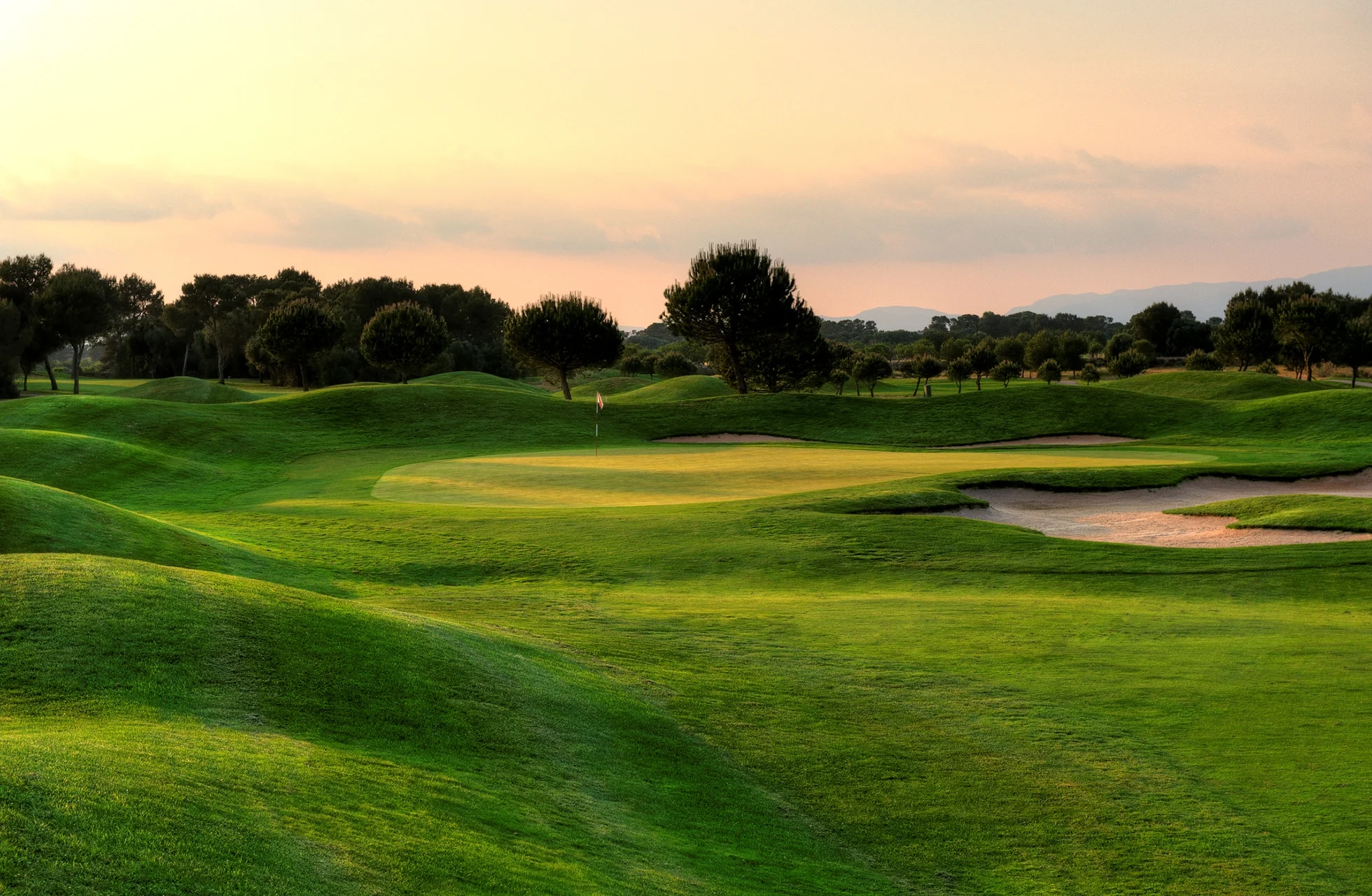 golfgreen på son antem golf med bunker i forgrunnen, omgitt av bølgende fairwayer og trær i varmt kveldslys