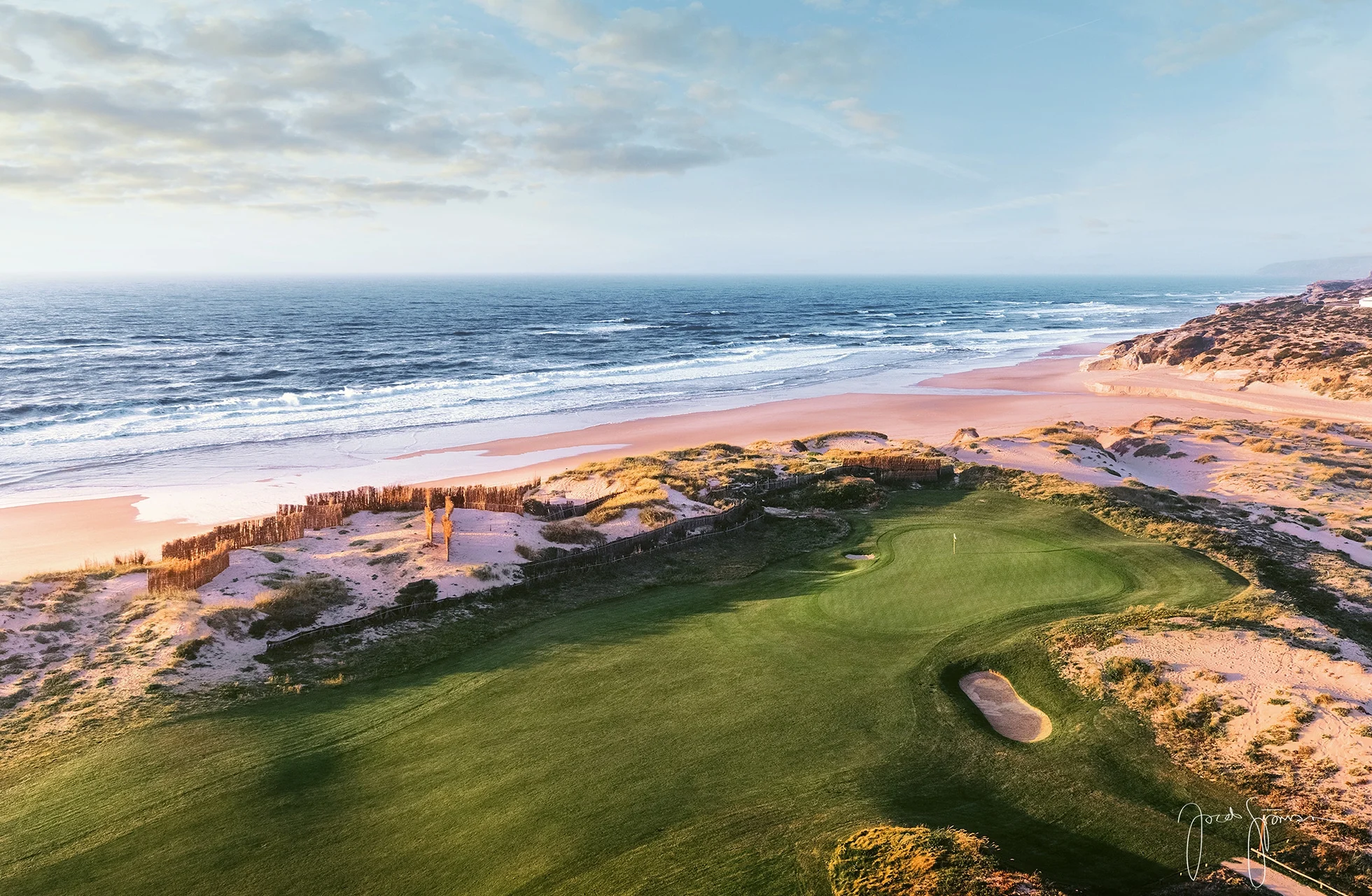 golfgreen med bunker i forgrunden, omgivet af naturlige klitter og kortklippede fairways, med strand og hav synligt i baggrunden i blødt aftenslys
