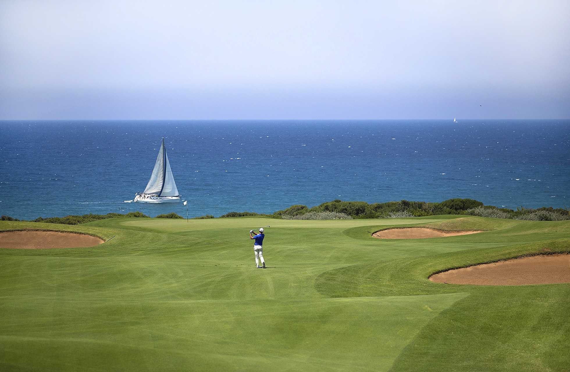 golfspiller slår slag på the dunes course, costa navarino, med bunkere og seilbåt på havet i bakgrunnen
