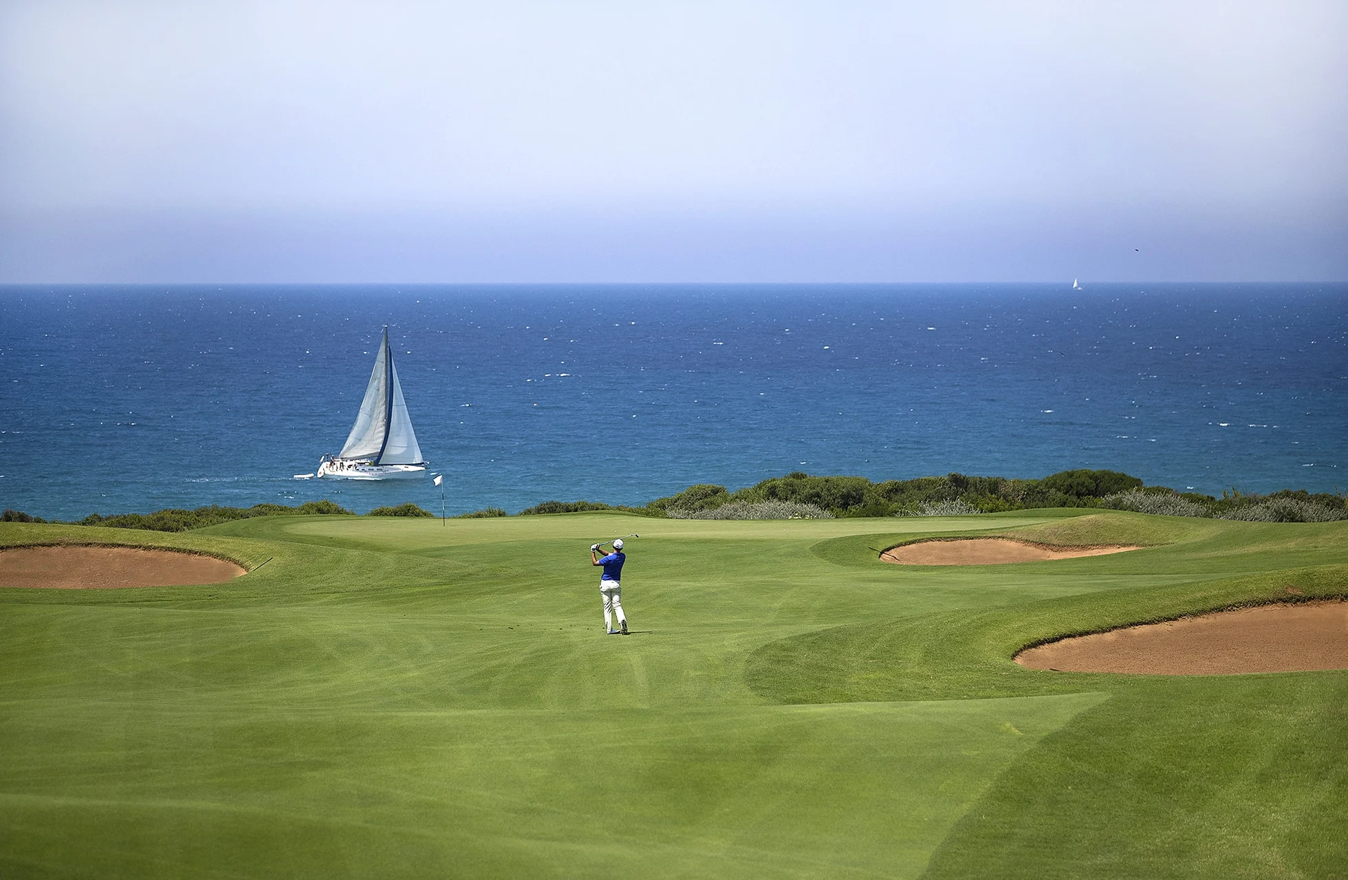 golfspiller slår slag på the dunes course, costa navarino, med bunkere og seilbåt på havet i bakgrunnen