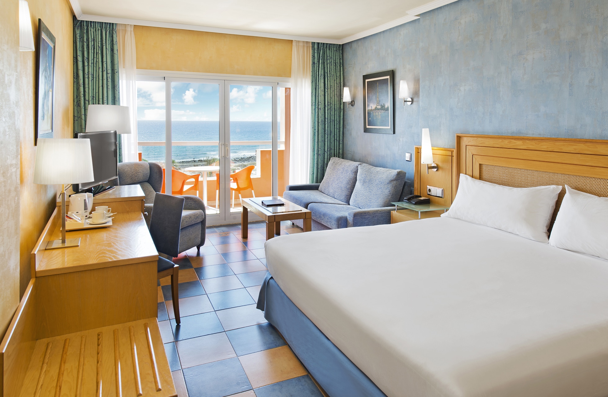 lyst og romslig hotellrom med dobbeltseng, skrivebord, sittegruppe og balkong med utemøbler og utsikt mot havet i dagslys