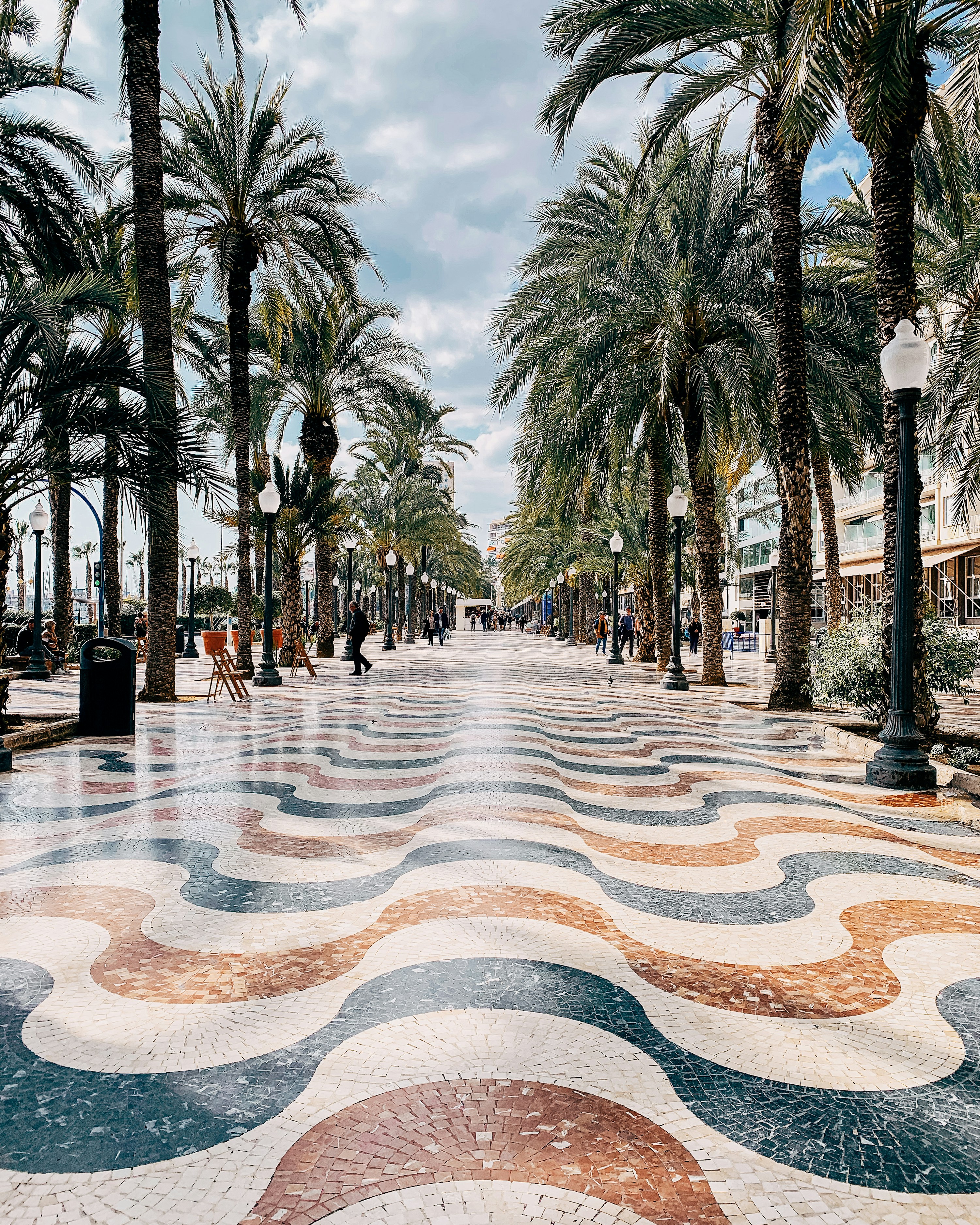 Palmekledd promenade med bølgeformet mosaikkgulv, gatelykter og mennesker som går langs den strandnære gangveien.
