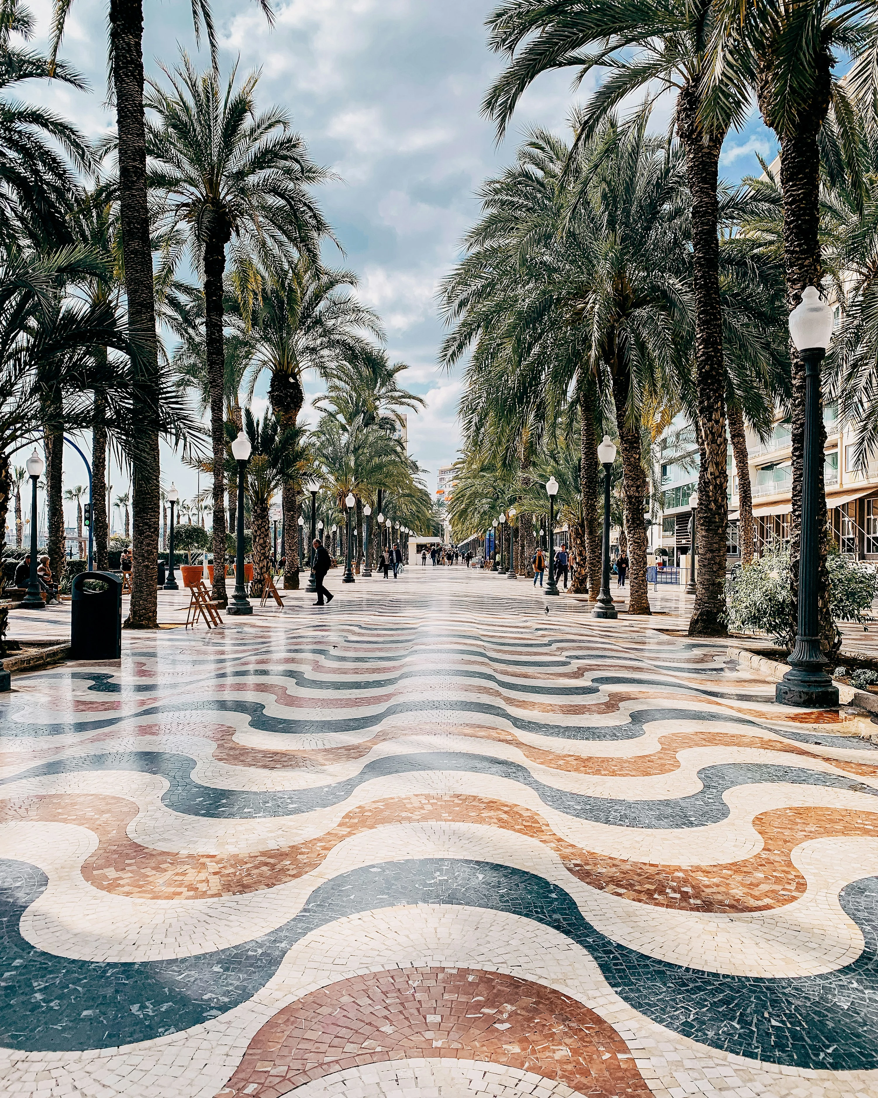 Palmekledd promenade med bølgeformet mosaikkgulv, gatelykter og mennesker som går langs den strandnære gangveien.
