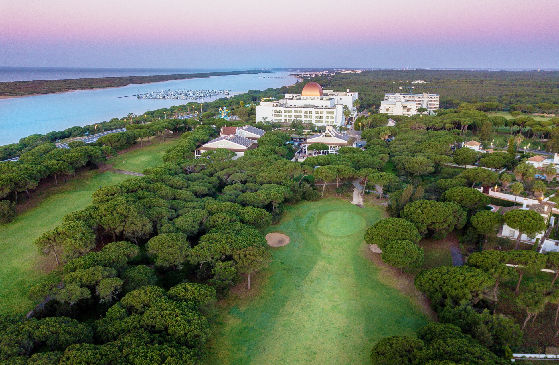 Luftfoto av stor hotellbygning ved golfbane nær kysten og lagunen, omgitt av furuskog.