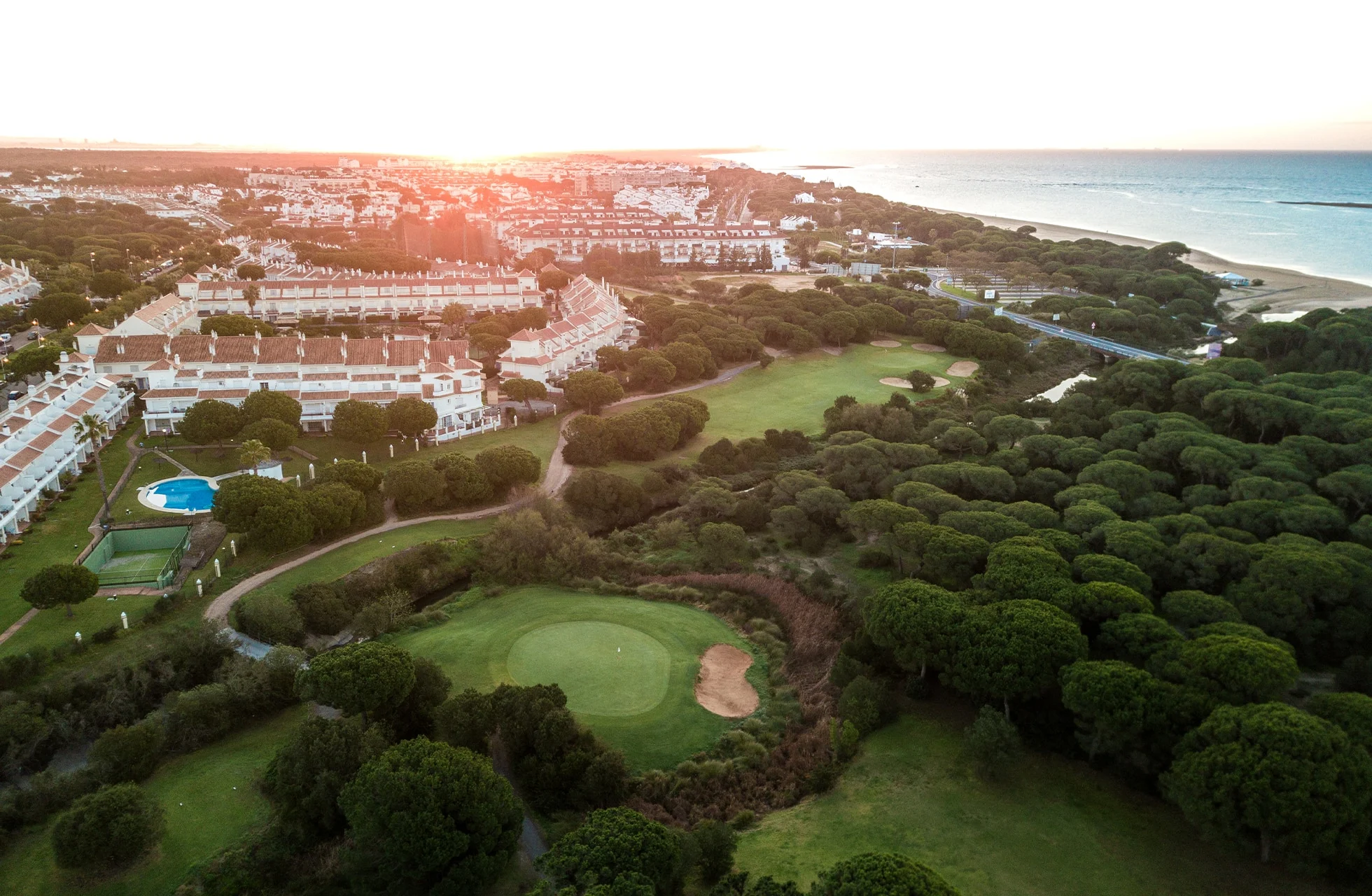 Droneutsikt over resortområde ved kysten i solnedgang, med golfbane og strand i bakgrunnen.
