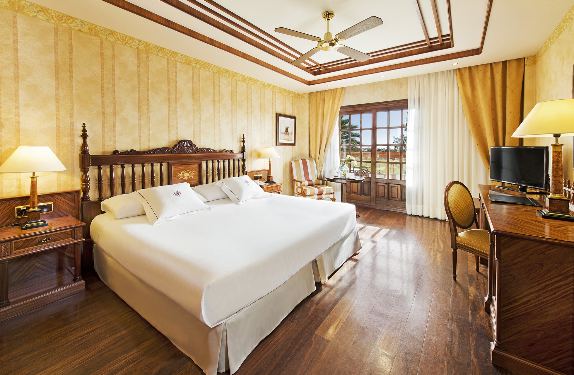 elegant hotellrom på elba palace golf med dobbeltseng, tregulv, skrivebord, takvifte og balkong med utemøbler i dagslys