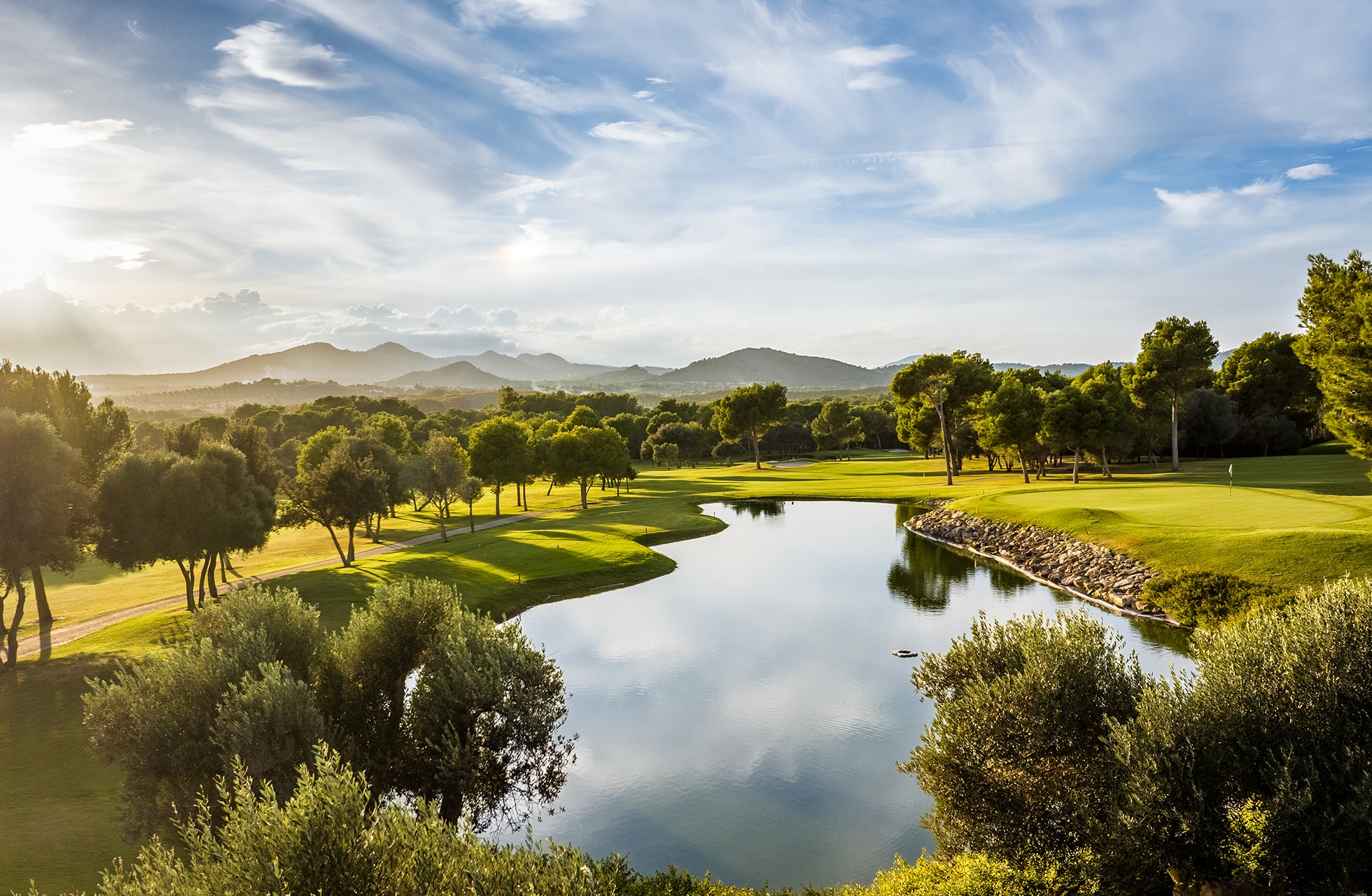 golfbane på son servera golf med stille vannhinder som speiler trær og himmel, omgitt av fairway, green og kupert landskap med fjell i bakgrunnen i dagslys