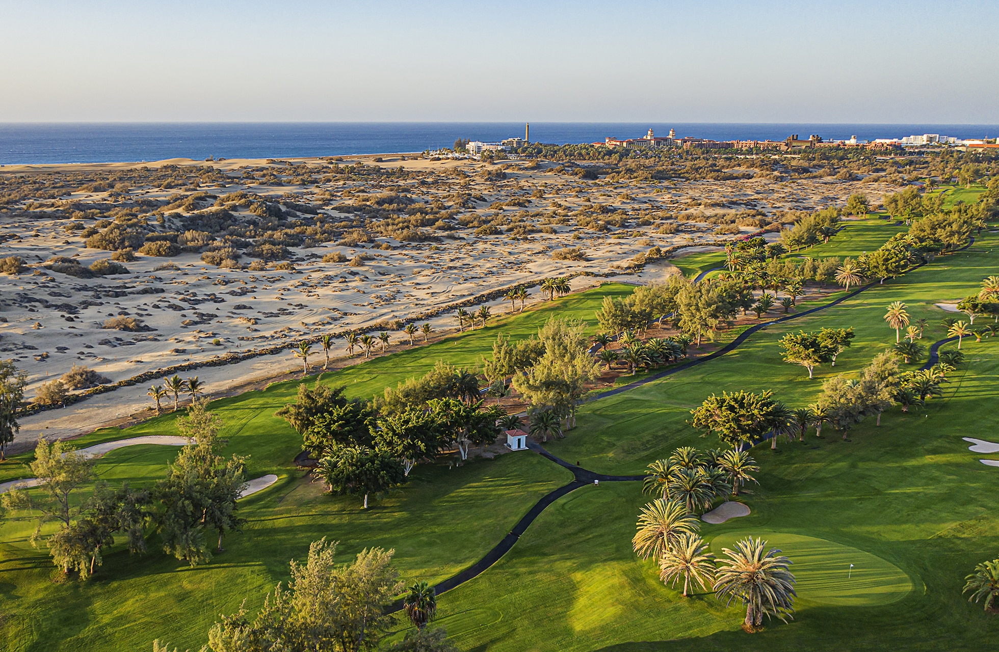 flyfoto av golfbane med grønn fairway og green i kontrast til sanddyner, med havet og kystlinjen i bakgrunnen i klart dagslys
