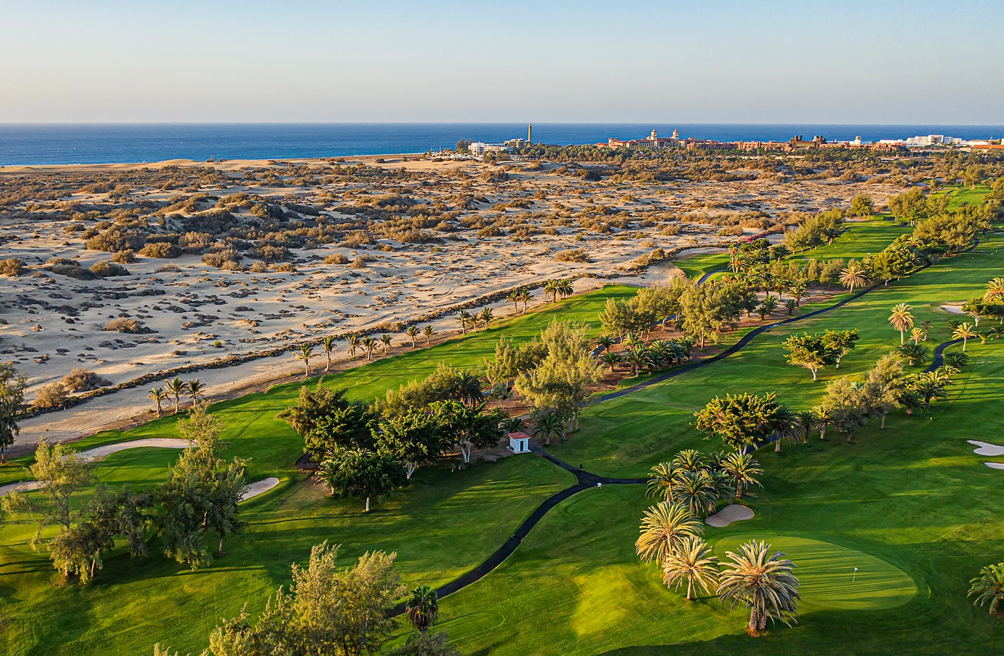 flyfoto av golfbane med grønn fairway og green i kontrast til sanddyner, med havet og kystlinjen i bakgrunnen i klart dagslys