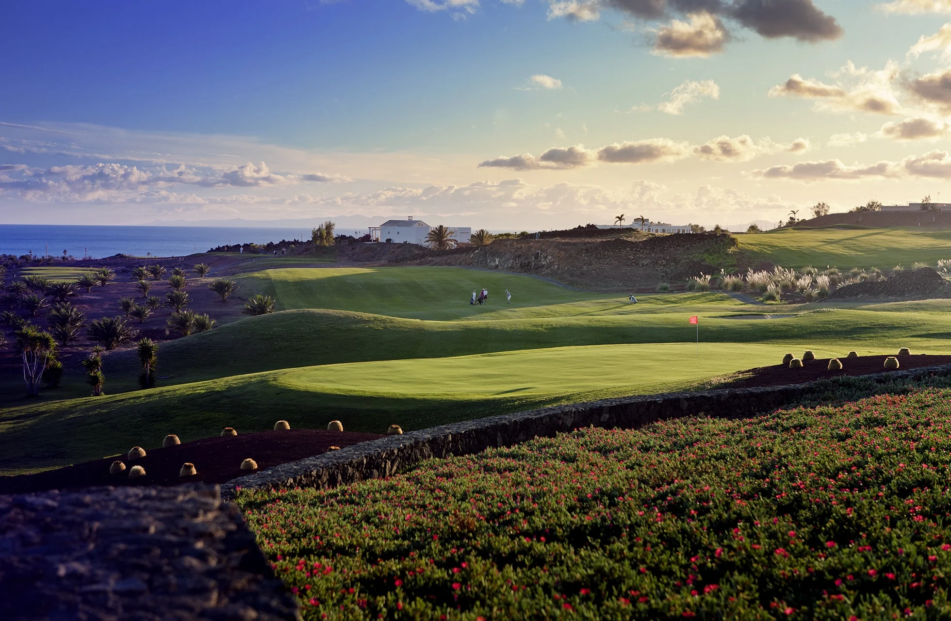 bølgende golfbane med green i forgrunnen og flagg på hullet. banen er omgitt av åpent landskap med lav vegetasjon, bygninger i det fjerne og havet ved horisonten under lys himmel.