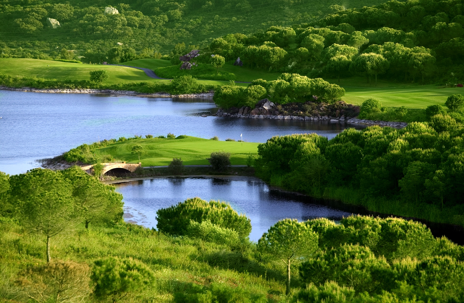 panoramautsikt over golfbane med green og fairway ved to vannpartier, forbundet med en liten bro i grønt og kupert landskap
