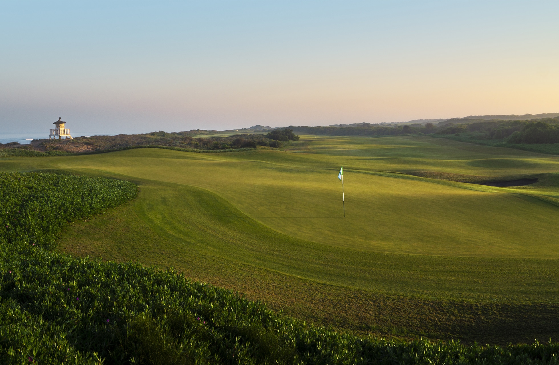 vidstrakt golfgreen med flagg i sentrum, omgitt av bølgende fairwayer og lav vegetasjon, med havet synlig i bakgrunnen i mykt kveldslys