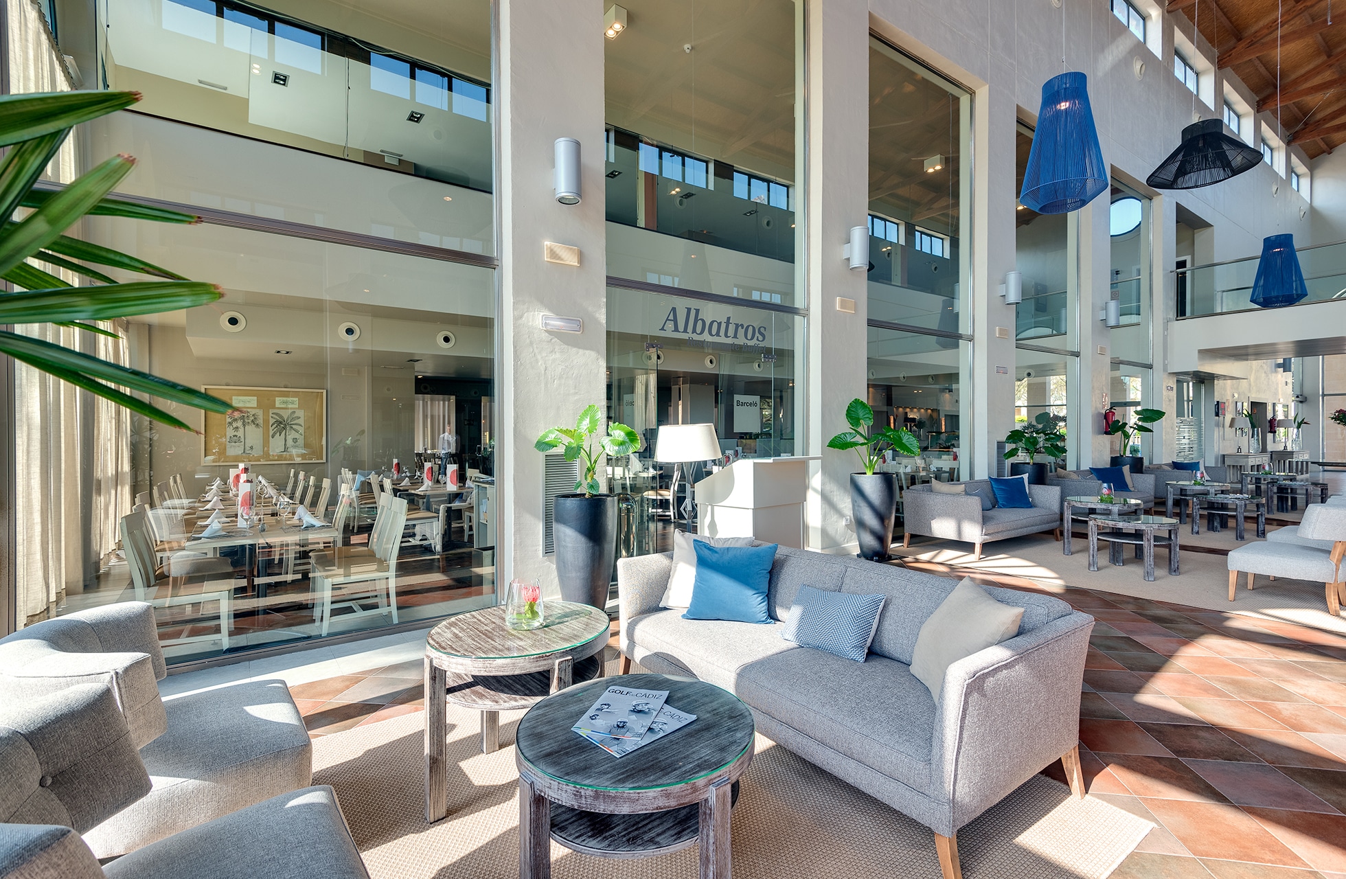 lys og romslig hotellobby med sofaer og lenestoler rundt små bord, store potteplanter, høyt under taket og glasspartier, med restaurantinngang og synlig skilt albatros i bakgrunnen i dagslys