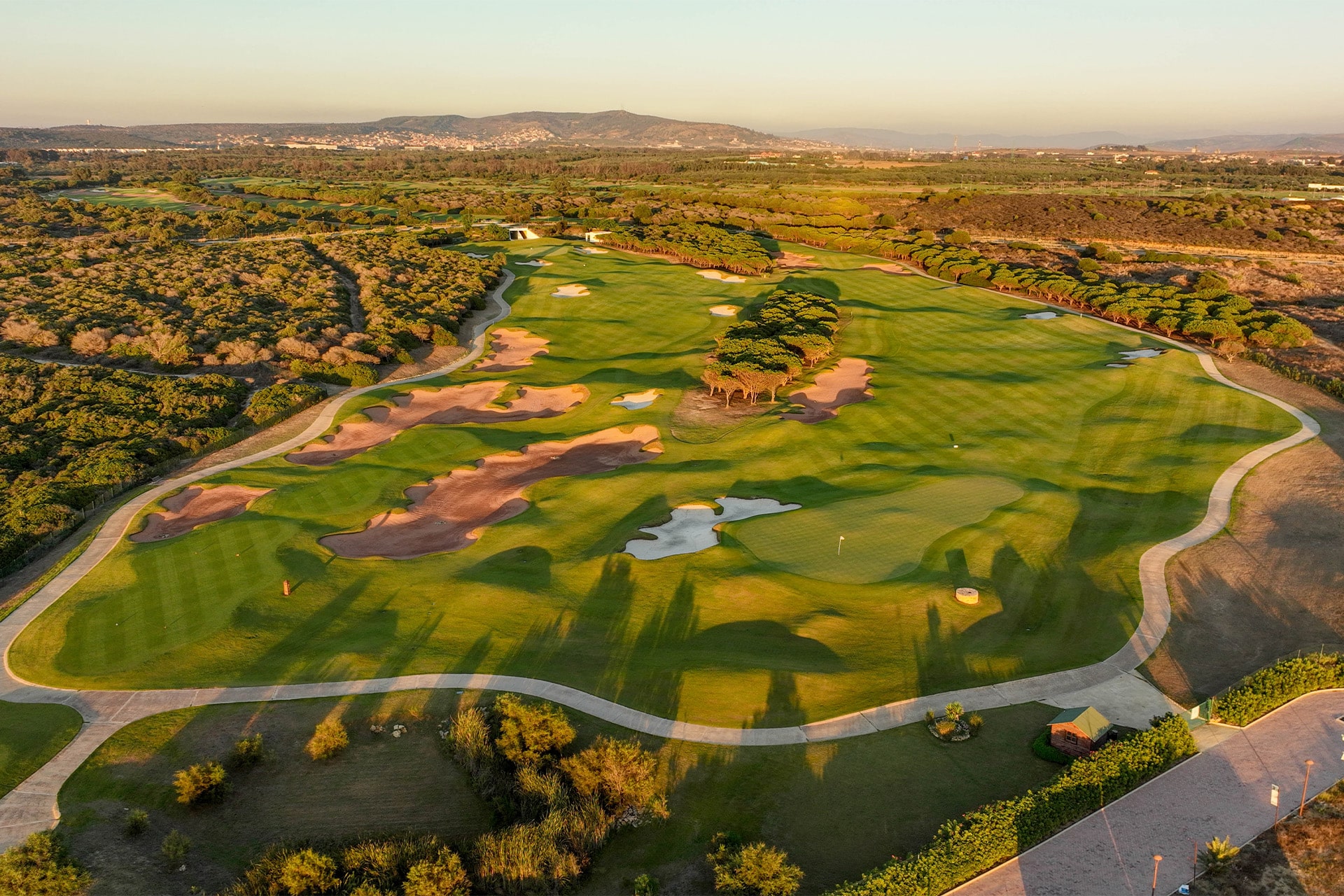 Luftfoto over en frodig golfbane med bølgende fairways, sandbunkers, vandhazarder og snoede golfveje i et åbent landskab i varmt aftenslys.
