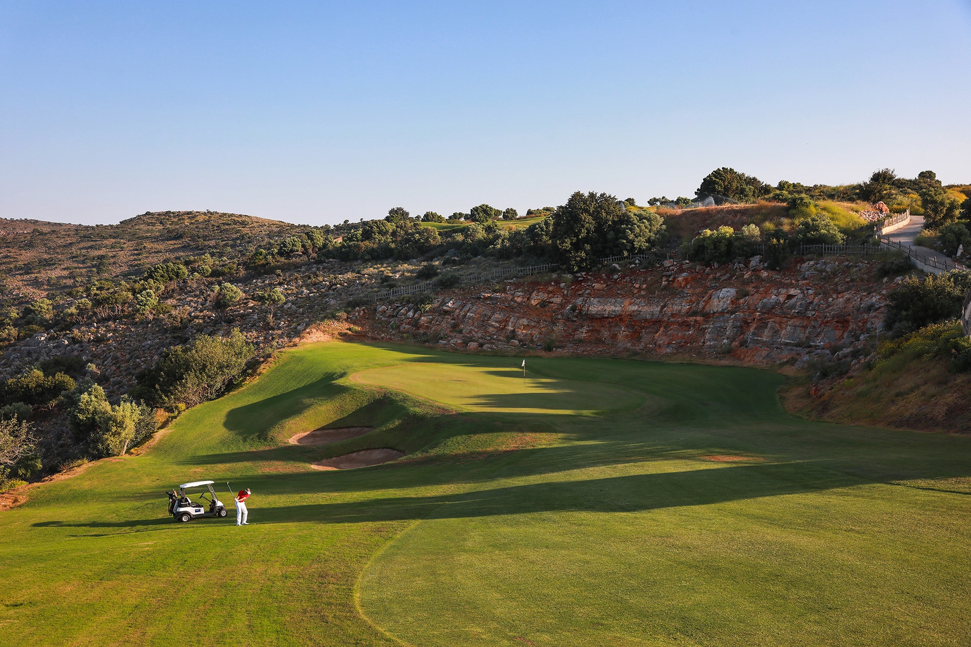 golfbane med green og bunkere i kupert landskap, en golfbil og en golfspiller ses i forgrunnen, omgitt av klipper og vegetasjon.