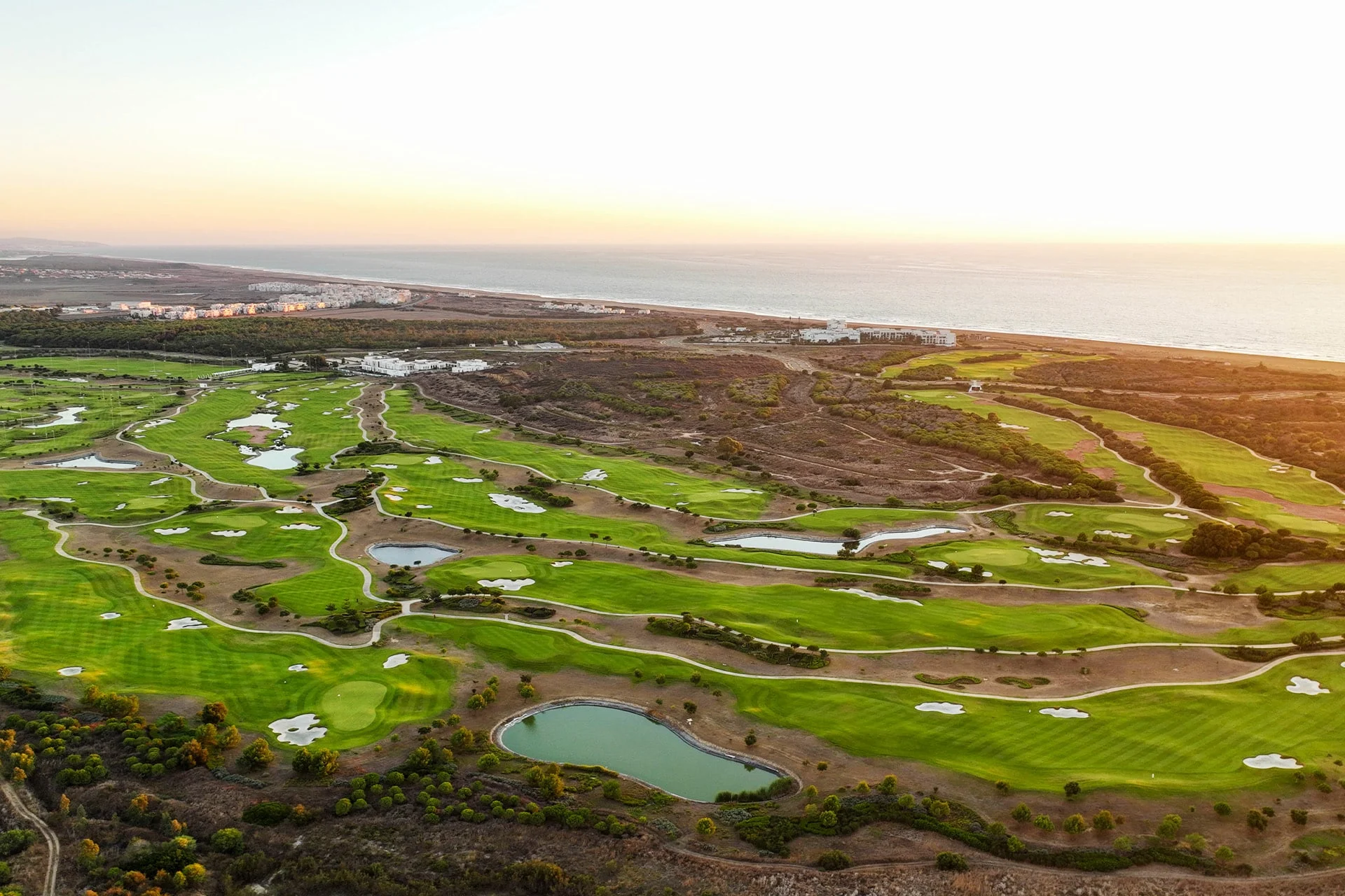 luftfoto af en vidstrakt golfbane med flere greens, fairways, bunkers og vandhazarder, omgivet af naturlig vegetation med havet synligt i baggrunden i varmt aftenslys