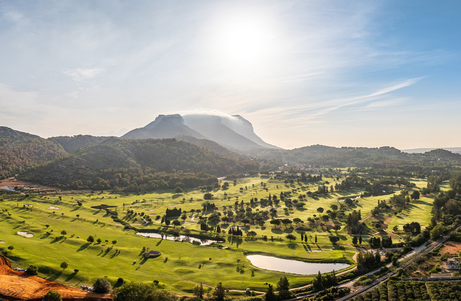 Panoramautsikt over golfbanen med fjell og sol i bakgrunnen.
