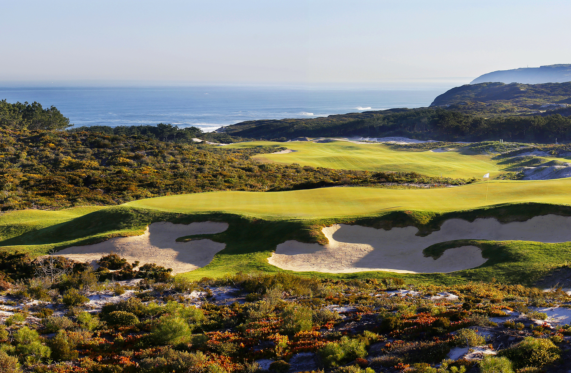 golfgreen med flere bunkere i forgrunnen, omgitt av naturlig vegetasjon og sanddyner, med havet og klippekyst synlig i bakgrunnen i klart dagslys