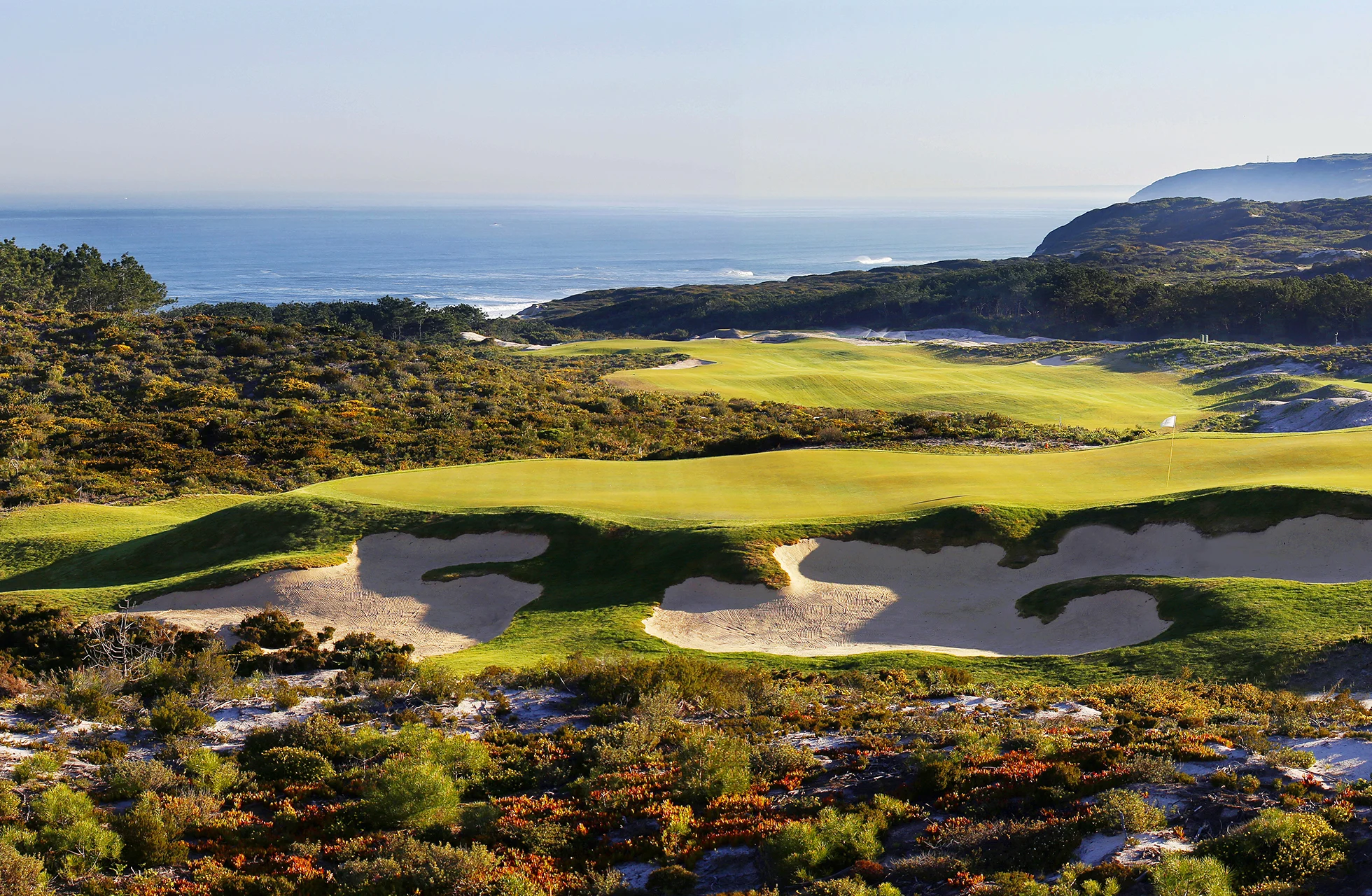 golfgreen med flere bunkere i forgrunnen, omgitt av naturlig vegetasjon og sanddyner, med havet og klippekyst synlig i bakgrunnen i klart dagslys