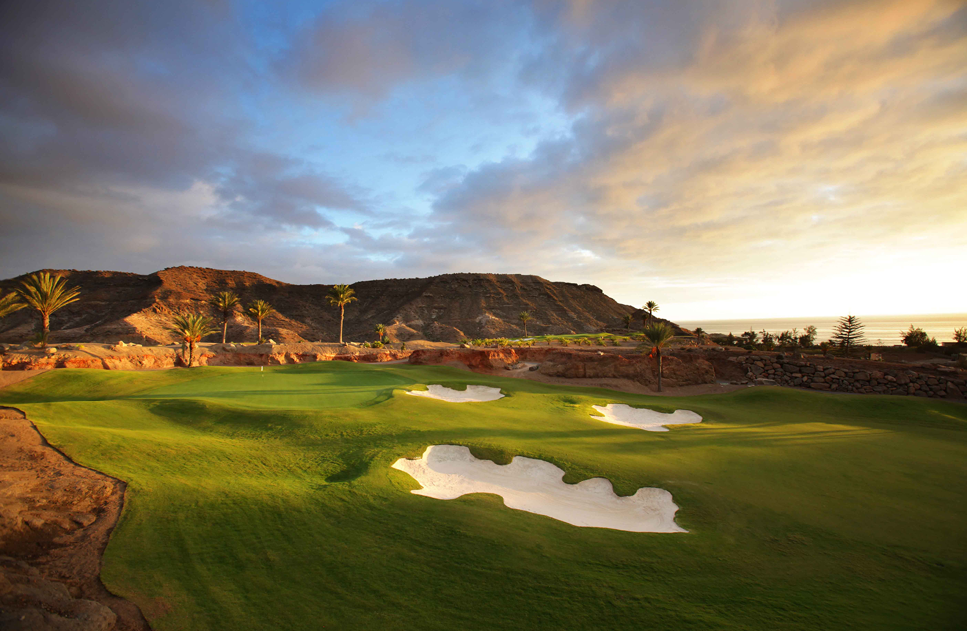 golfgreen på anfi tauro golf med flere bunkere i kupert landskap, grønne gressflater og fjell i bakgrunnen i varmt kveldslys
