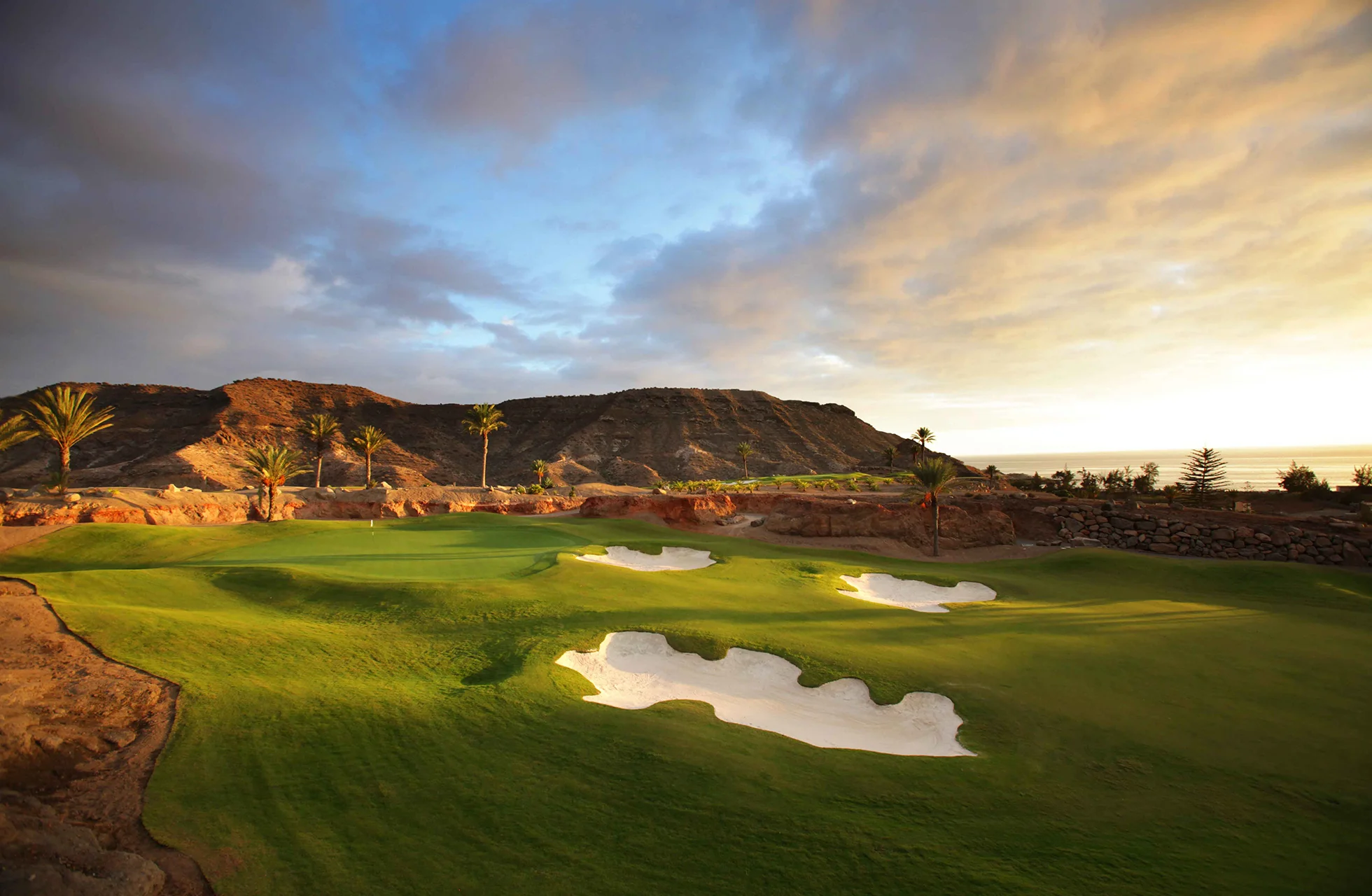 golfgreen på anfi tauro golf med flere bunkere i kupert landskap, grønne gressflater og fjell i bakgrunnen i varmt kveldslys