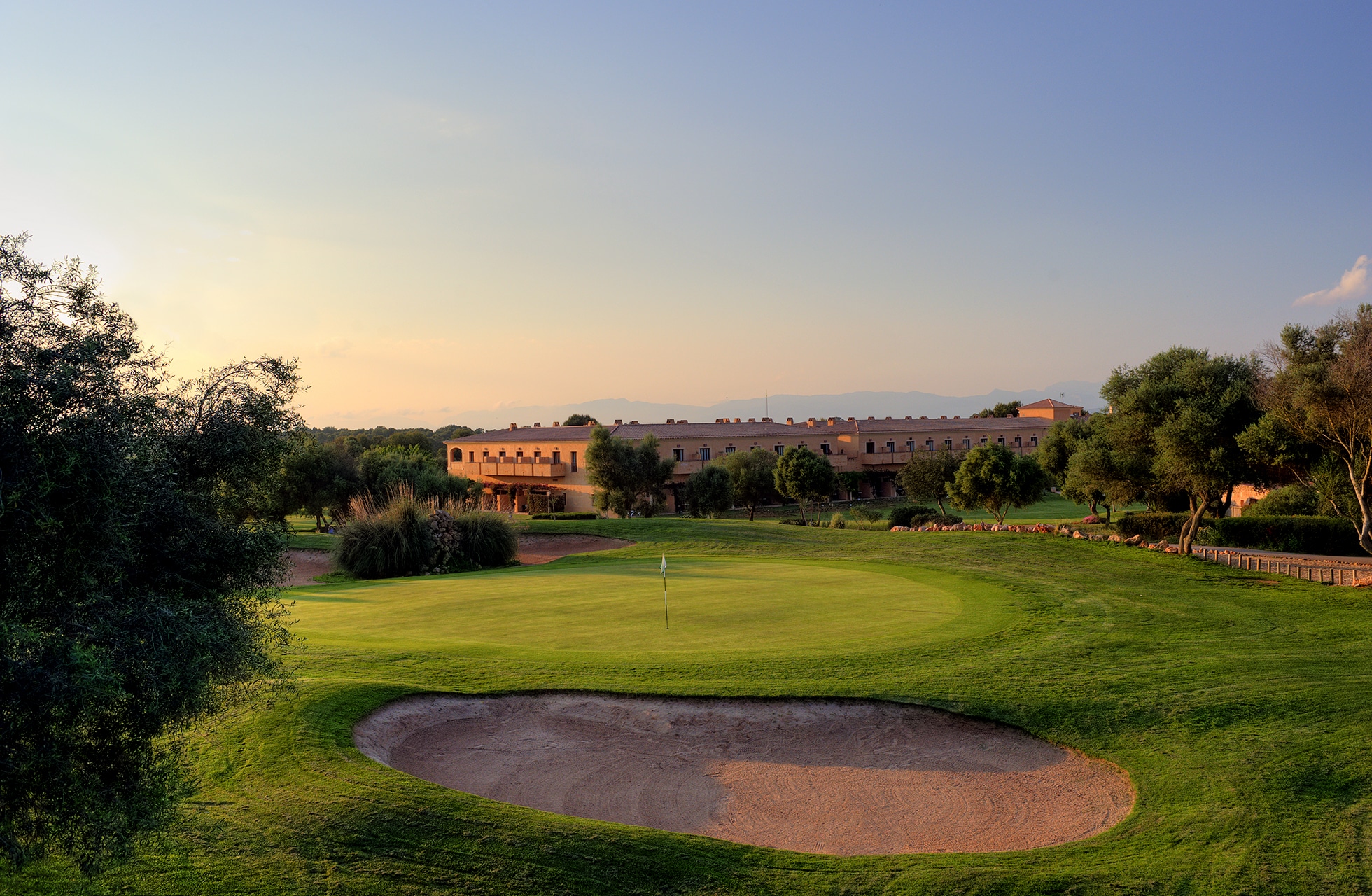 golfgreen med sandbunker i forgrunnen, flagg på greenen og bygninger i bakgrunnen i varmt kveldslys.