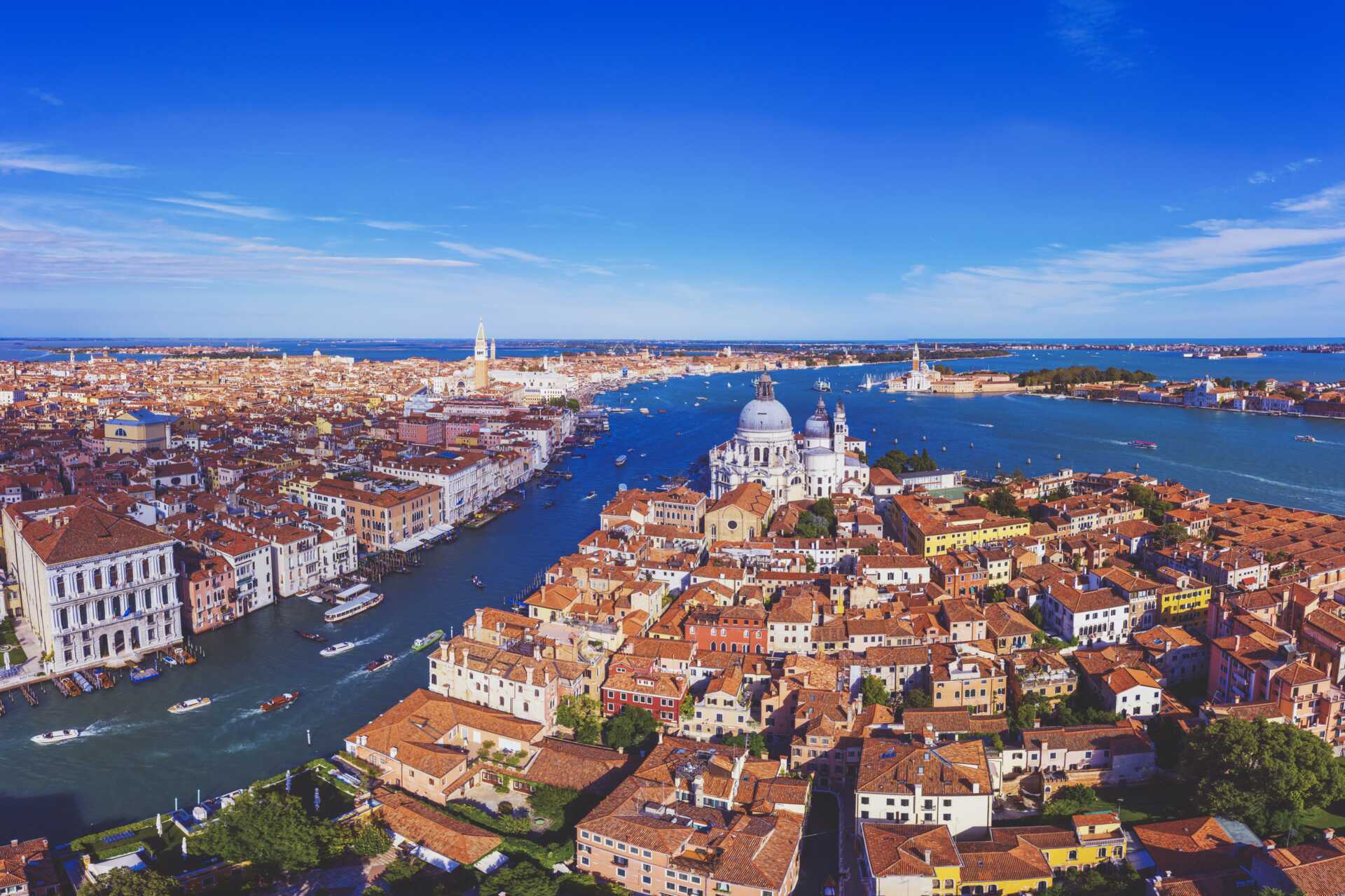 ilmakuva venetsiasta, jossa näkyvät canal grande, historialliset terrakotankattoiset rakennukset ja santa maria della saluten kirkko sinisen taivaan alla.