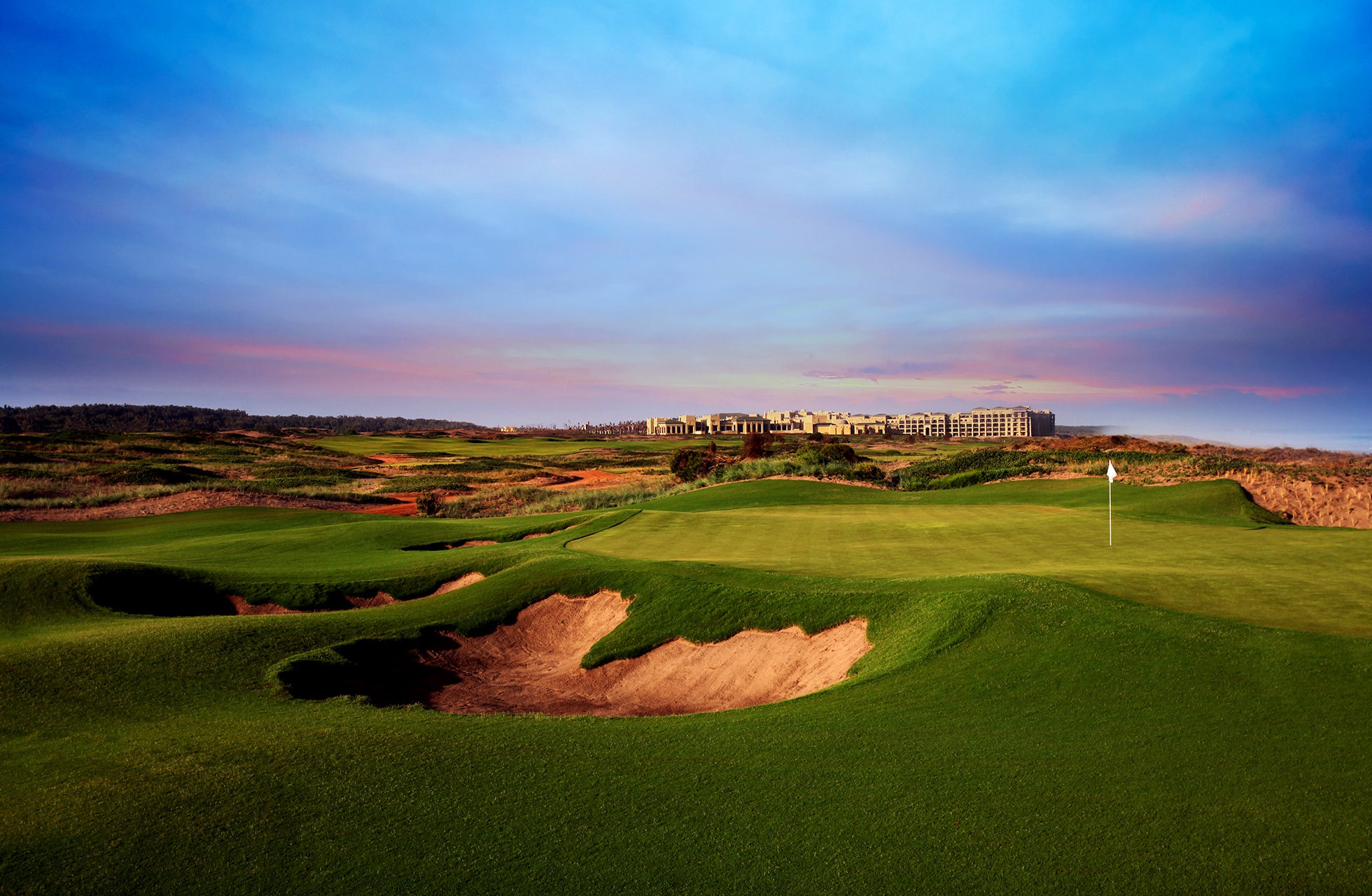 golfgreen med flagg og flere bunkere i kupert kystlandskap, grønne fairwayer og sandkontraster, bygninger i bakgrunnen under fargerik himmel