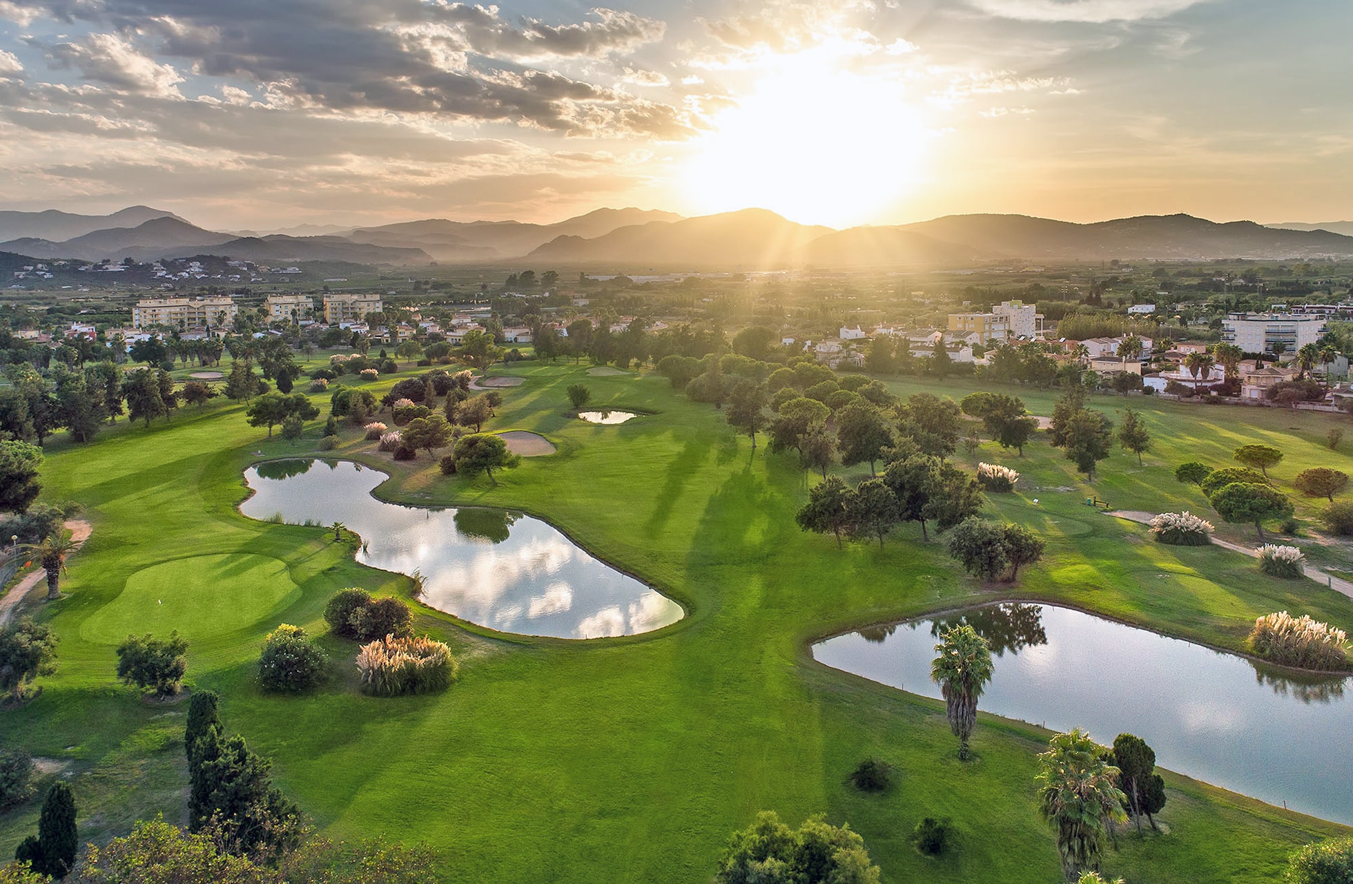 flyfoto av en grønn golfbane med flere vannhindre, bølgende fairwayer og trær, omgitt av bebyggelse, med solen lavt over horisonten i varmt kveldslys
